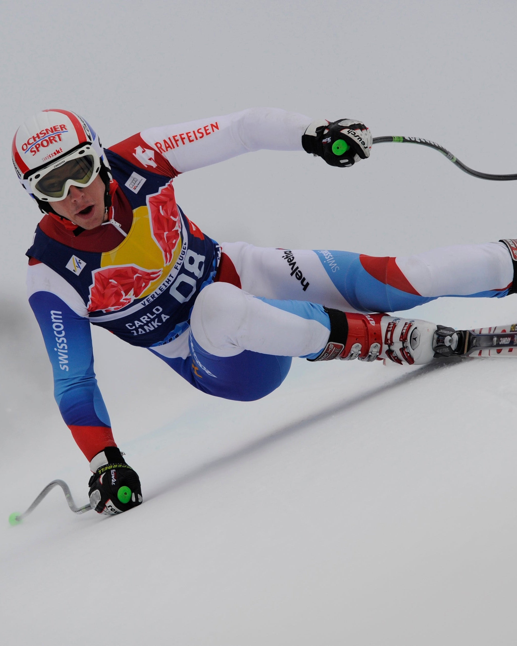 Carlo Janko in a dynamic side-angle pose during a race, wearing a Swiss team livery, a white helmet, and ski boots against a bright, overexposed snowy background.
