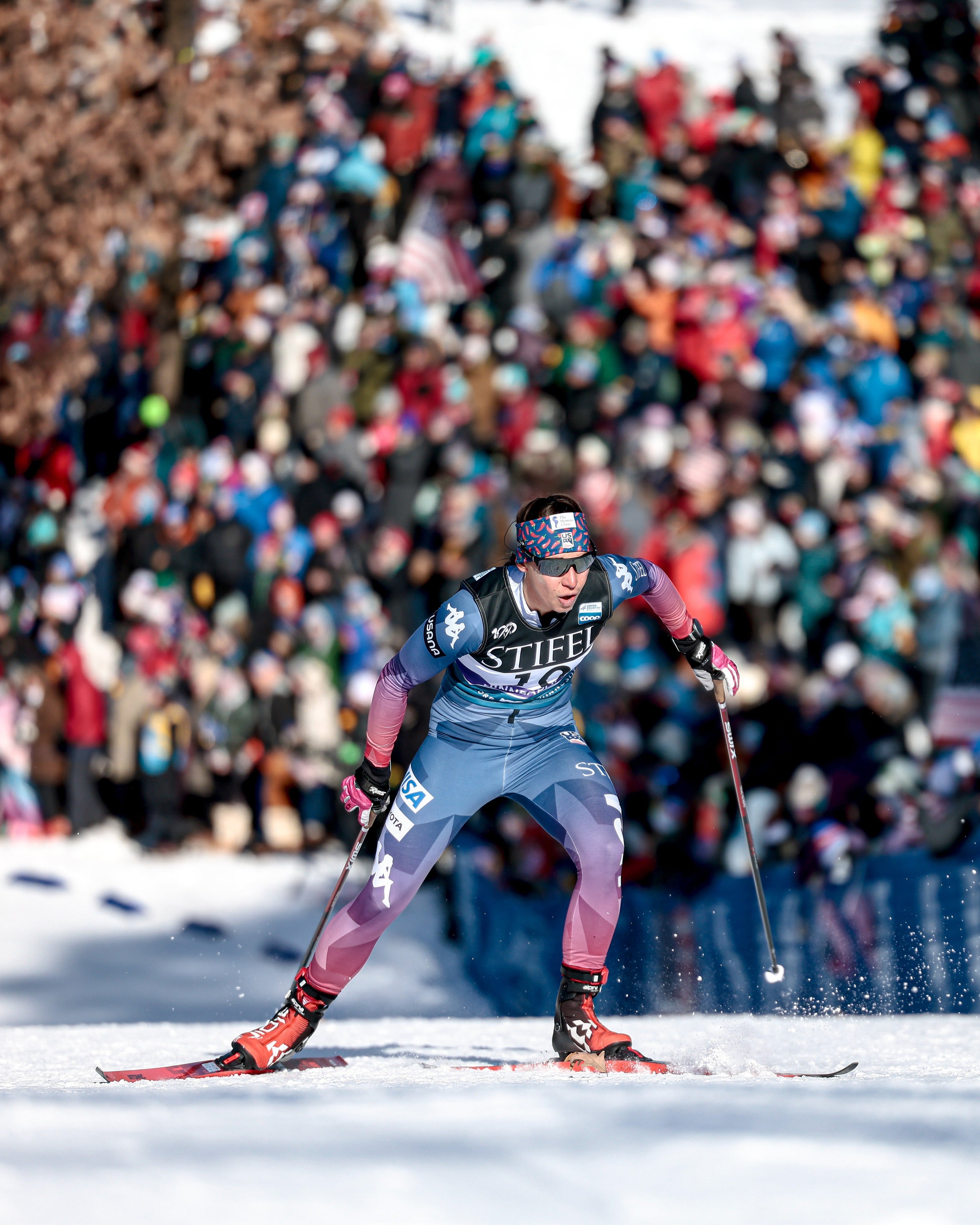 Julia Kern in a blue and purple ski suit, racing on a snowy track with a large blurred crowd in the background.