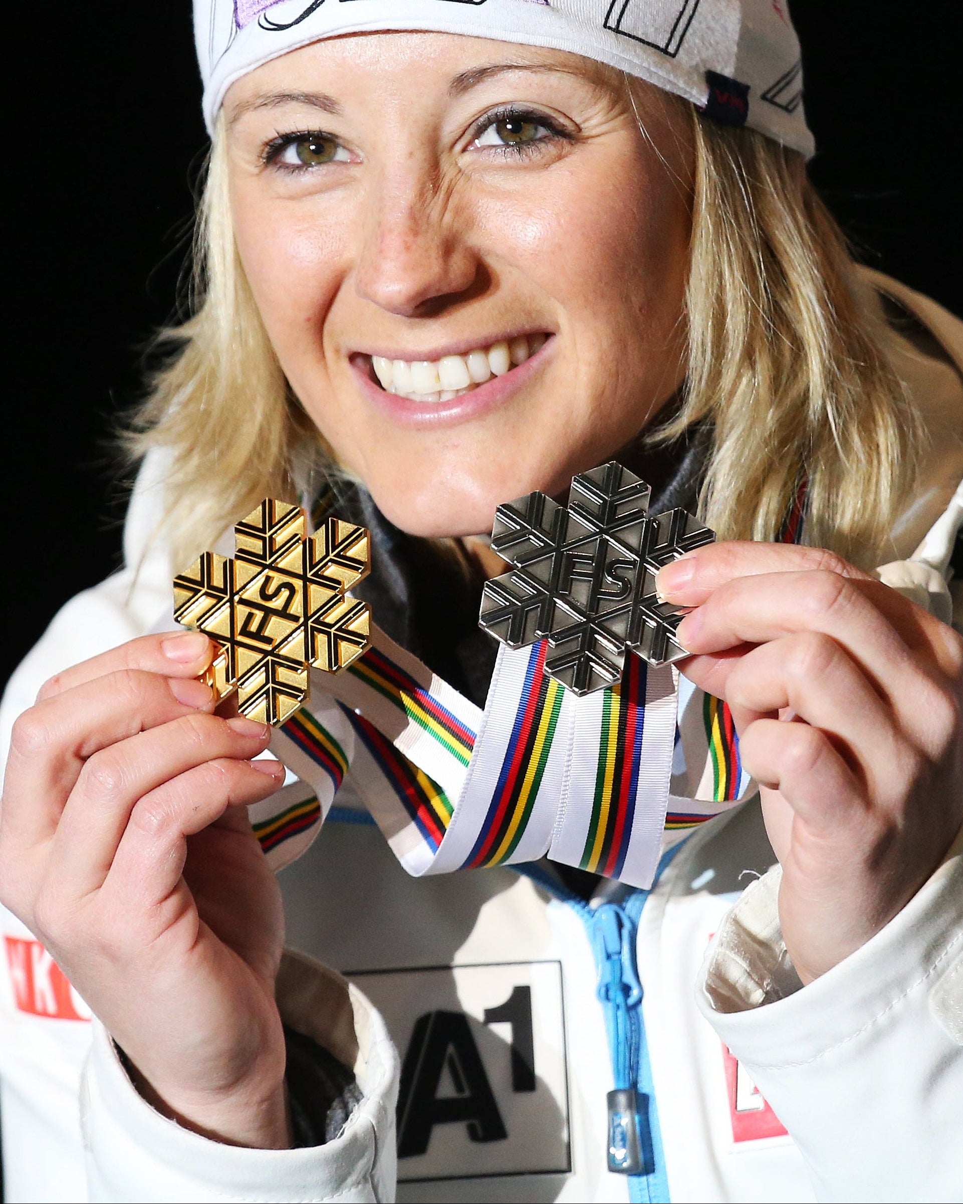 A smiley Michaela Kirchgasser in a white ski jacket and a white headband with a purple Milka logo holds up two medals with colorful ribbons against a black background.
