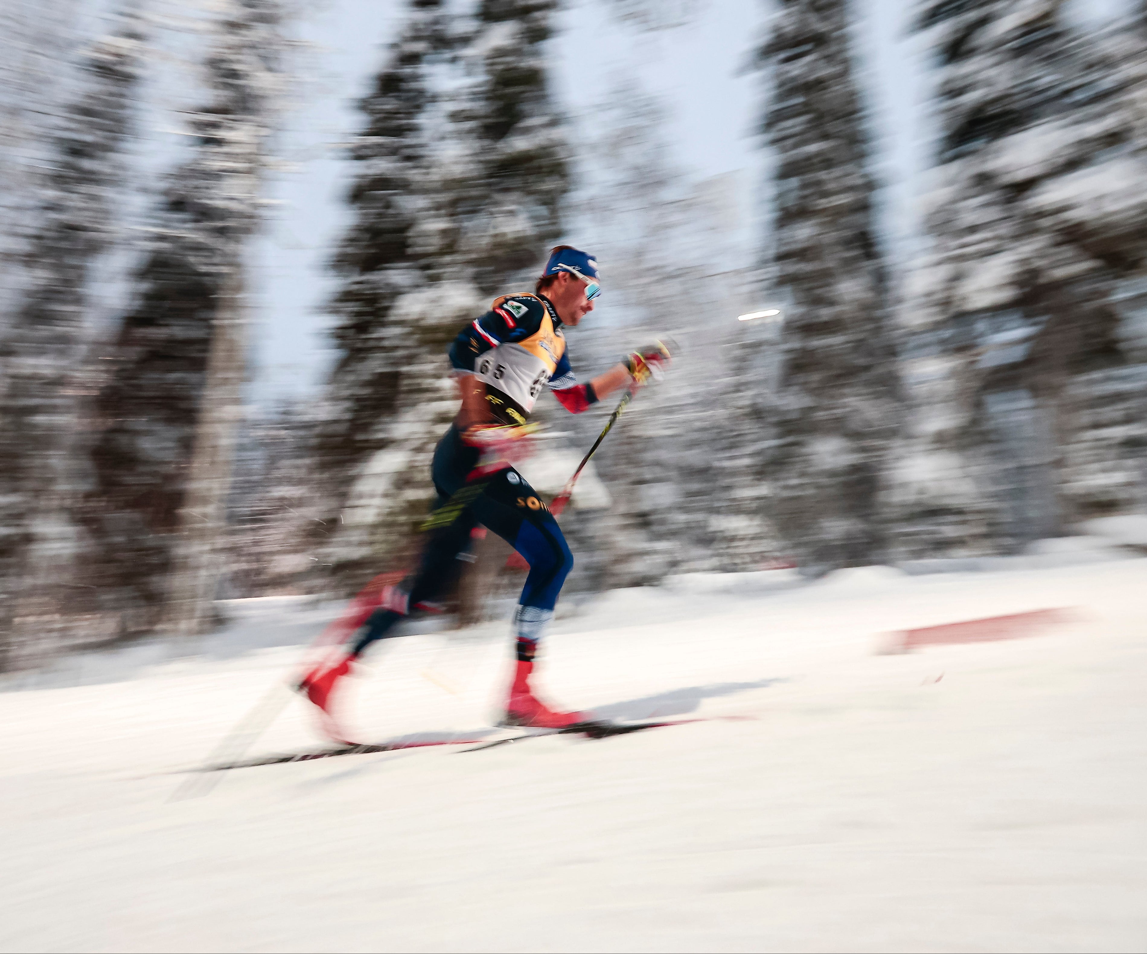 Hugo Lapalus in a blue and red outfit gliding through deep snow with blurred evergreen trees in the background, captured with a dynamic motion blur effect.