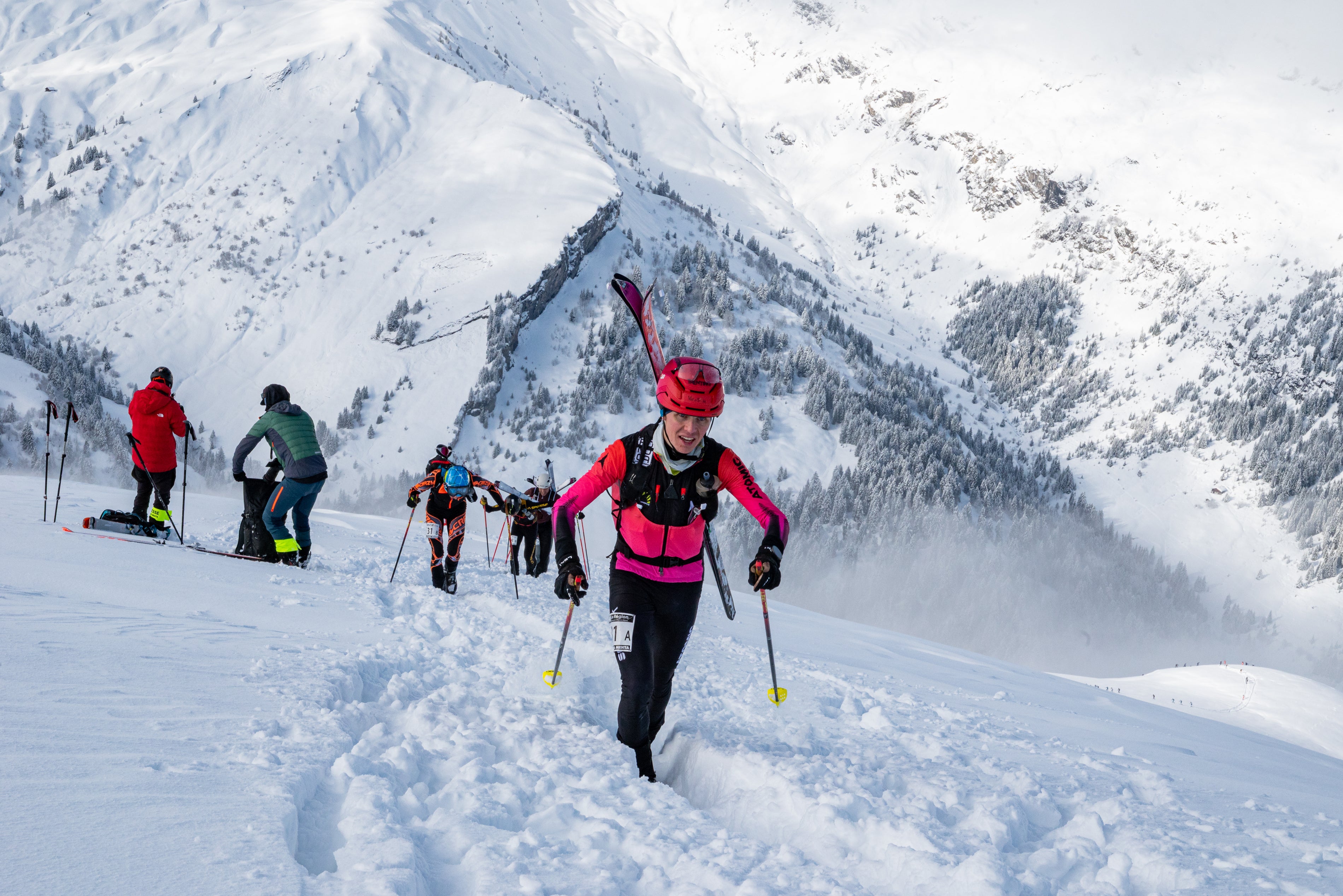 Davide Magnini in a bright pink and black jacket walks through deep snow on a mountain slope, carrying skis and using ski poles, with other skiers and snow-covered peaks in the background.
