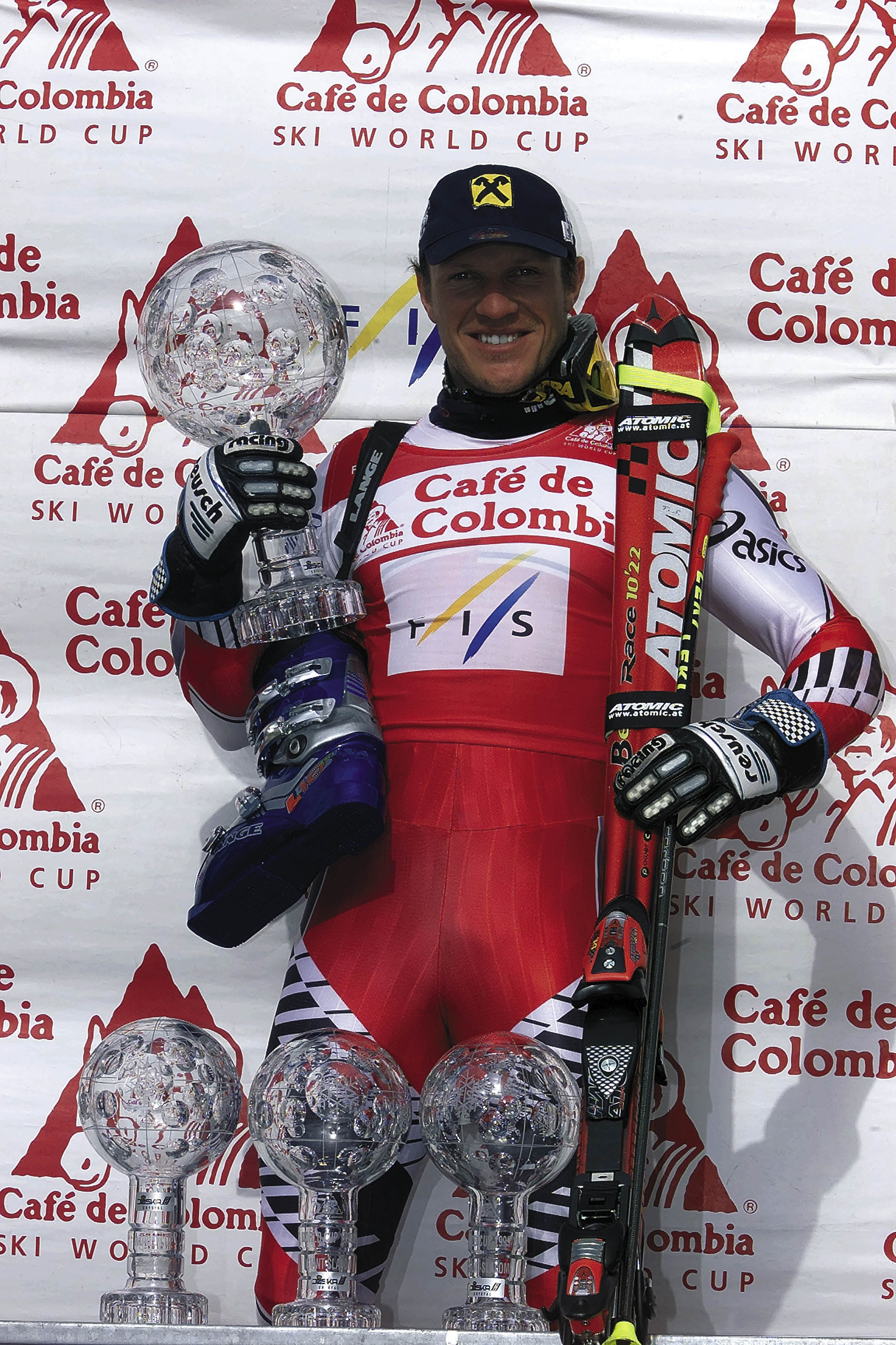 A smiley Hermann Maier stands on a podium holding a large silver trophy, with skis and additional trophies in front. The skier wears a red and white racing suit.