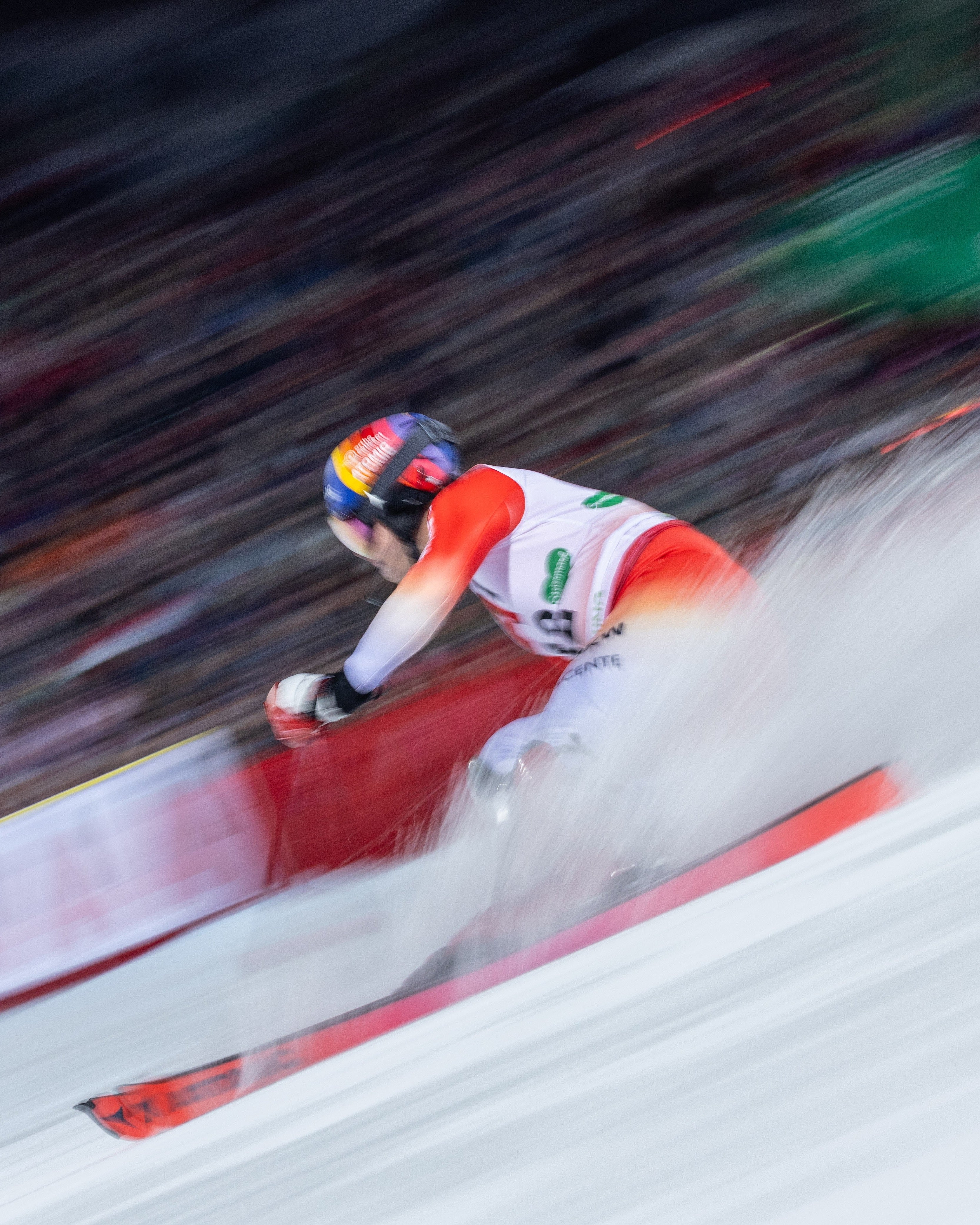 Tanguy Nef in a white, red racing suit carving through snow with a large spray of powder behind them, captured in a dynamic motion shot.