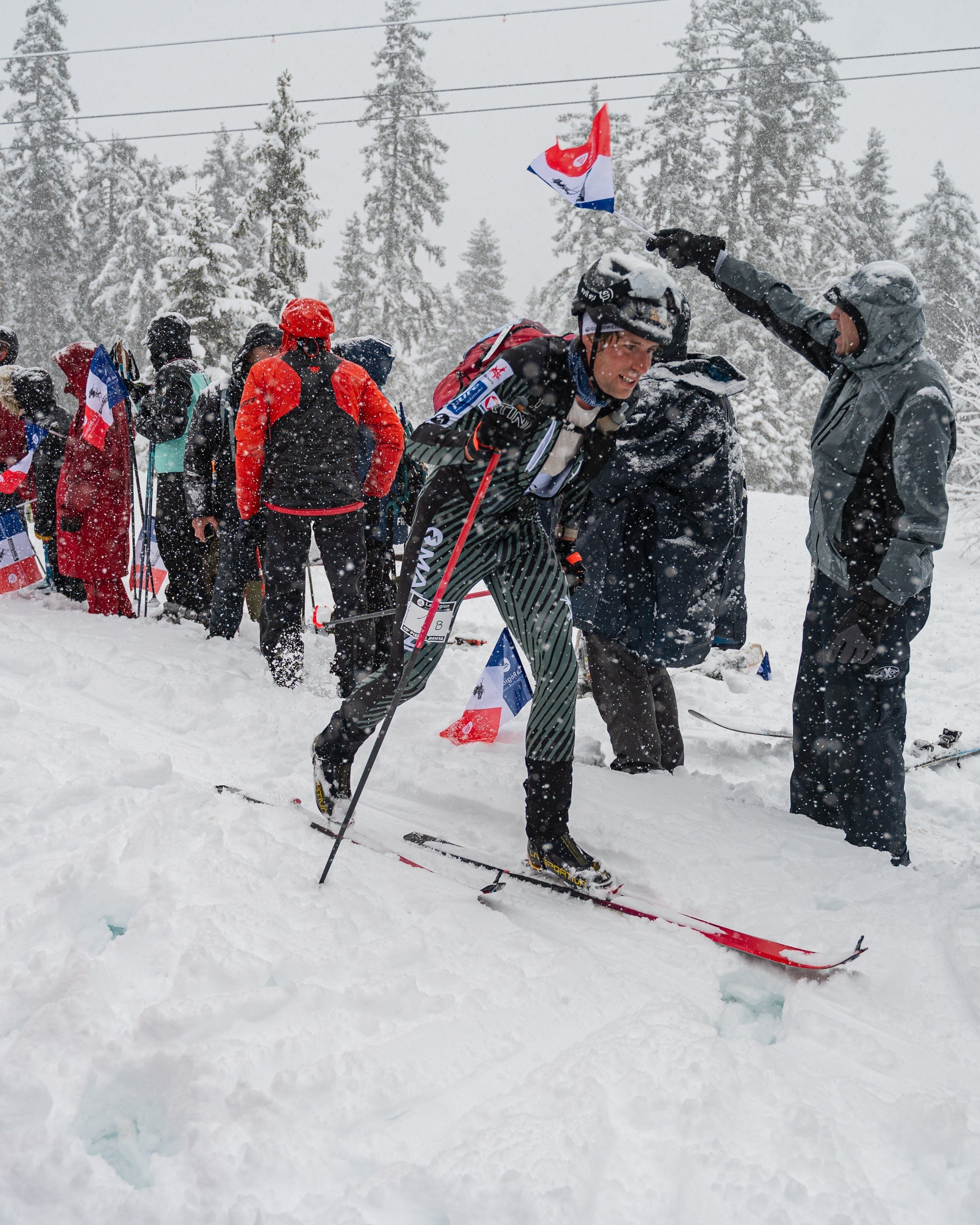 A group of fans cheering for Nils Oberauer on a race day.  Snow-covered pine trees and power lines are visible in the background. 