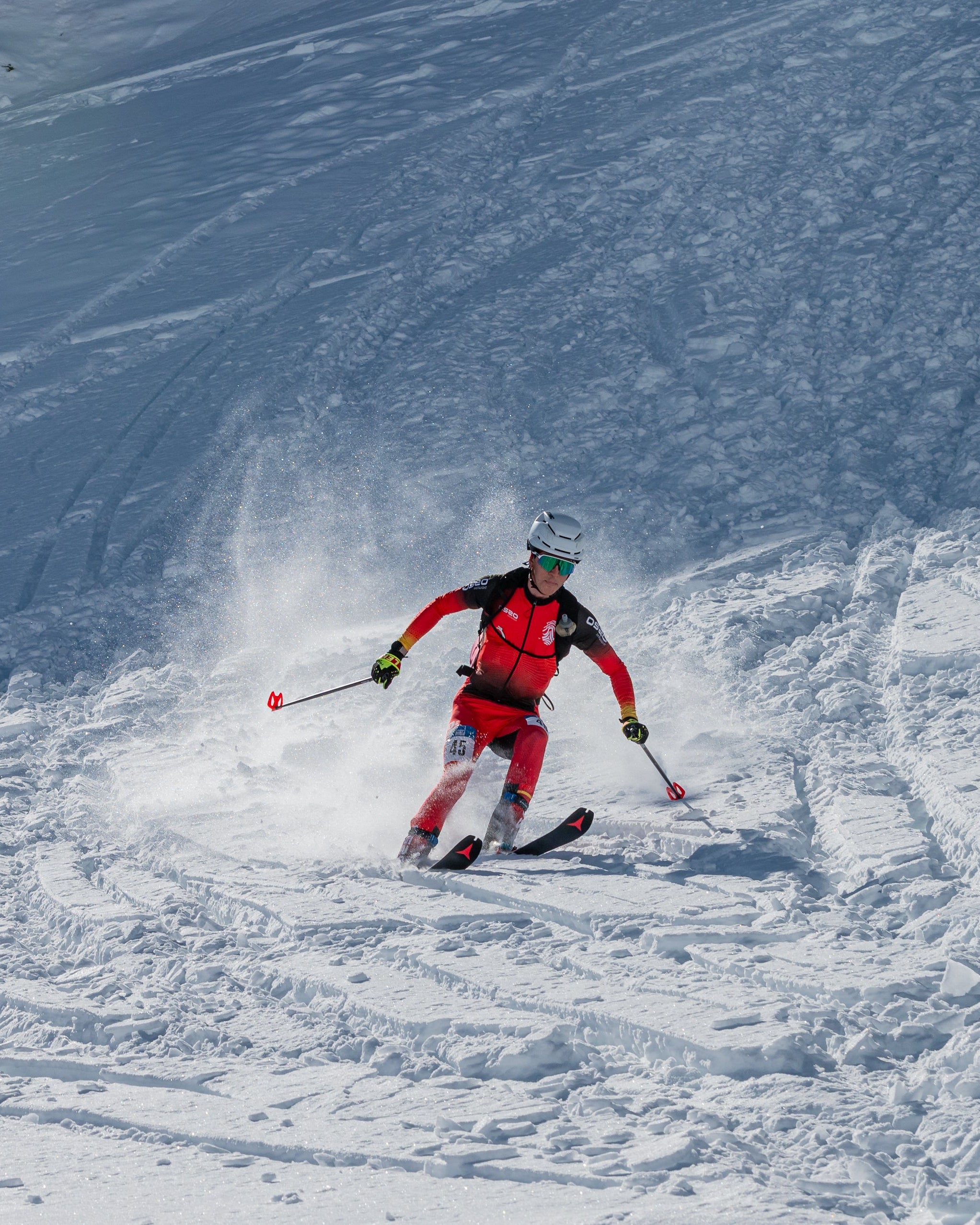 Albert Pérez in a red and black ski suit carving through fresh powder on a snowy mountain slope, with ski tracks and spray visible in the snow.