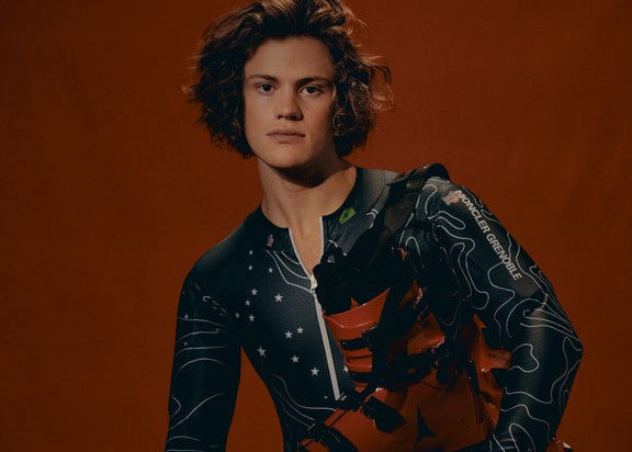 Lucas Pinheiro Braathen wearing a dark patterned racing suit, holding his ski boots, seated on a stool against a solid orange-brown studio background.