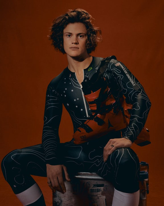 Lucas Pinheiro Braathen wearing a dark patterned racing suit, holding his ski boots, seated on a stool against a solid orange-brown studio background.