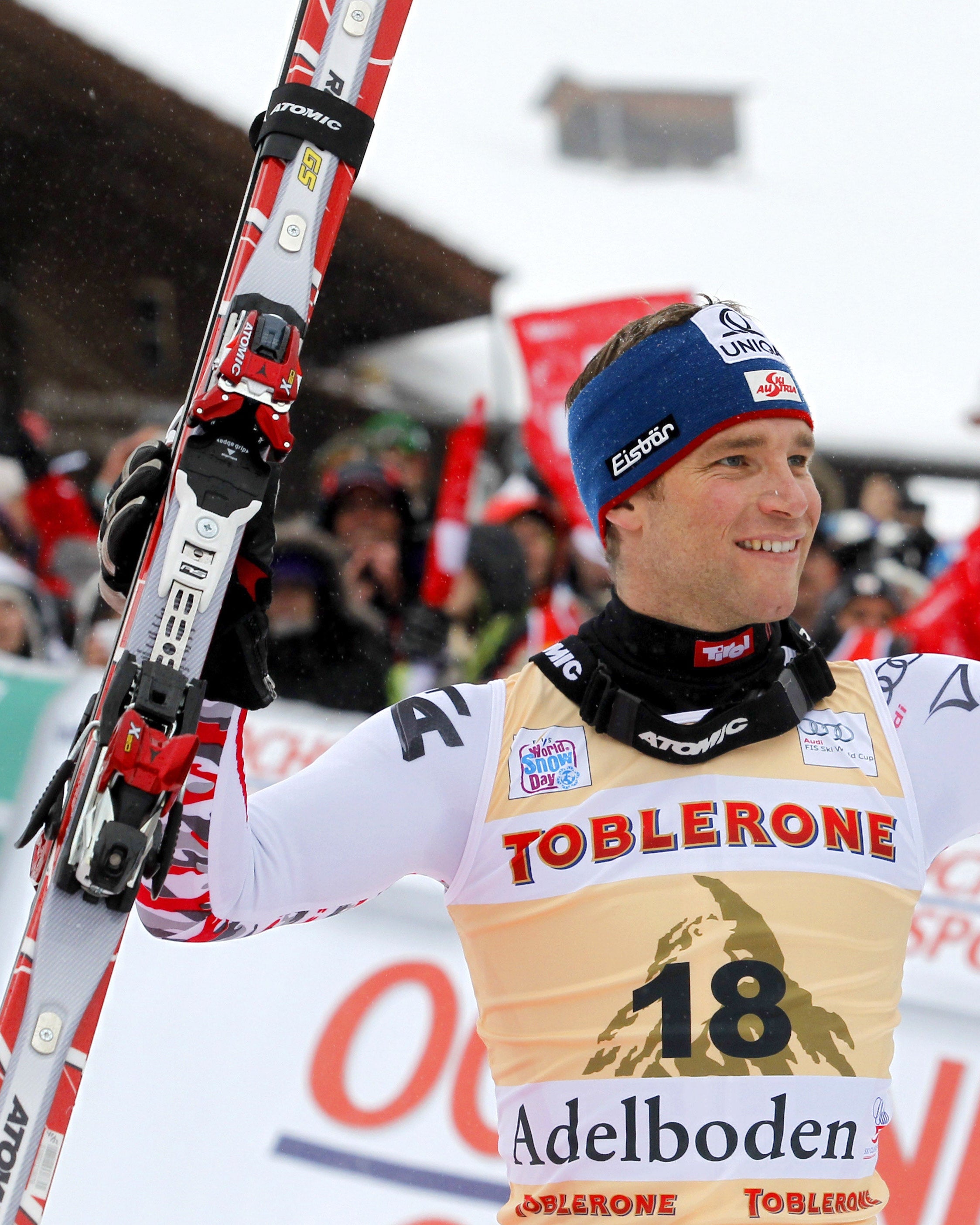 Benjamin Raich in a white racing suit with bib number 18, holding red and white skis and ski poles, celebrating on a snowy outdoor race course with spectators in the background.