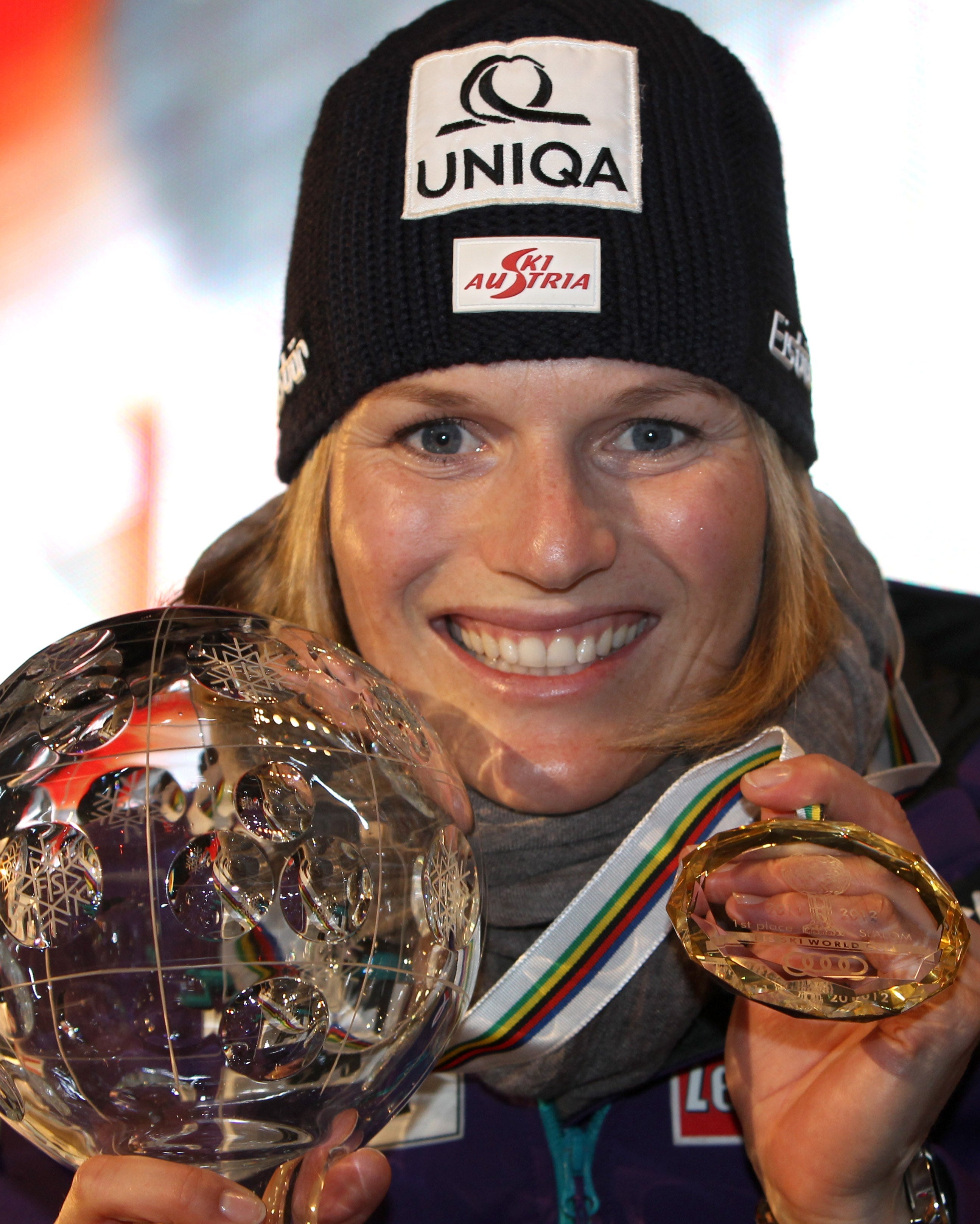 A smiley Marlies Raich in winter sports gear holds a large crystal trophy and a gold medal around her neck. She wears a black branded beanie and a purple jacket with sponsor patches.
