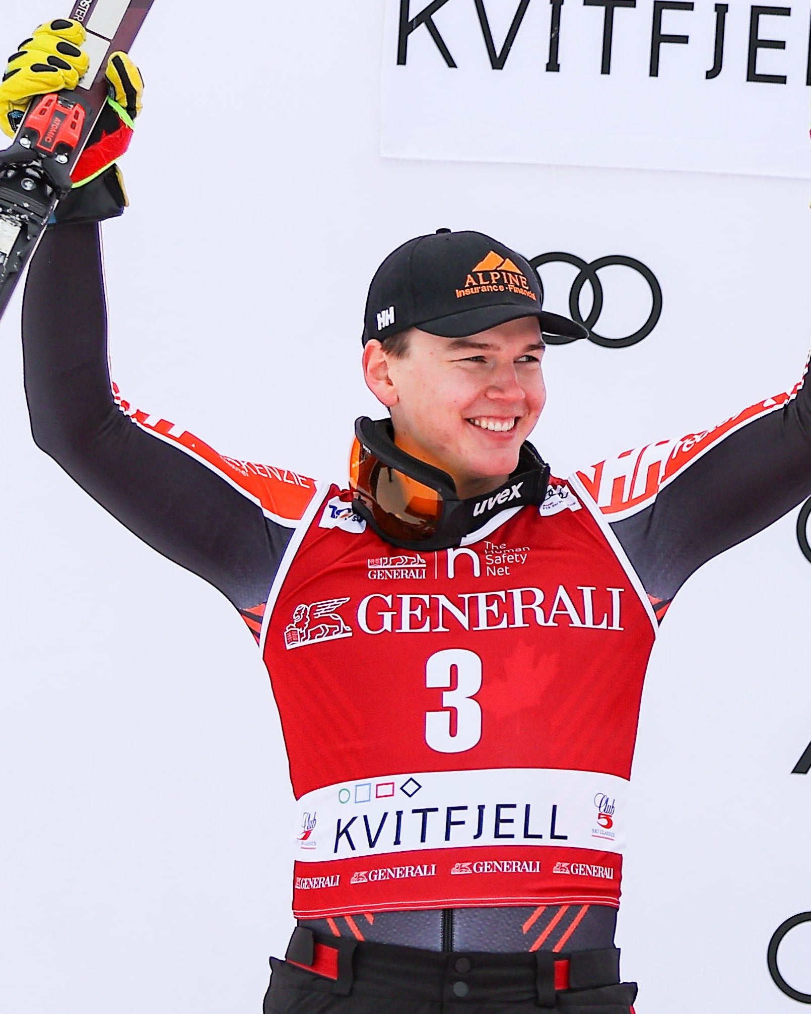 Jeffrey Read celebrates on a podium with her arms raised, holding skis and ski poles. He wears a red and black race bib with the number 3 and sponsor logos, along with a black cap.