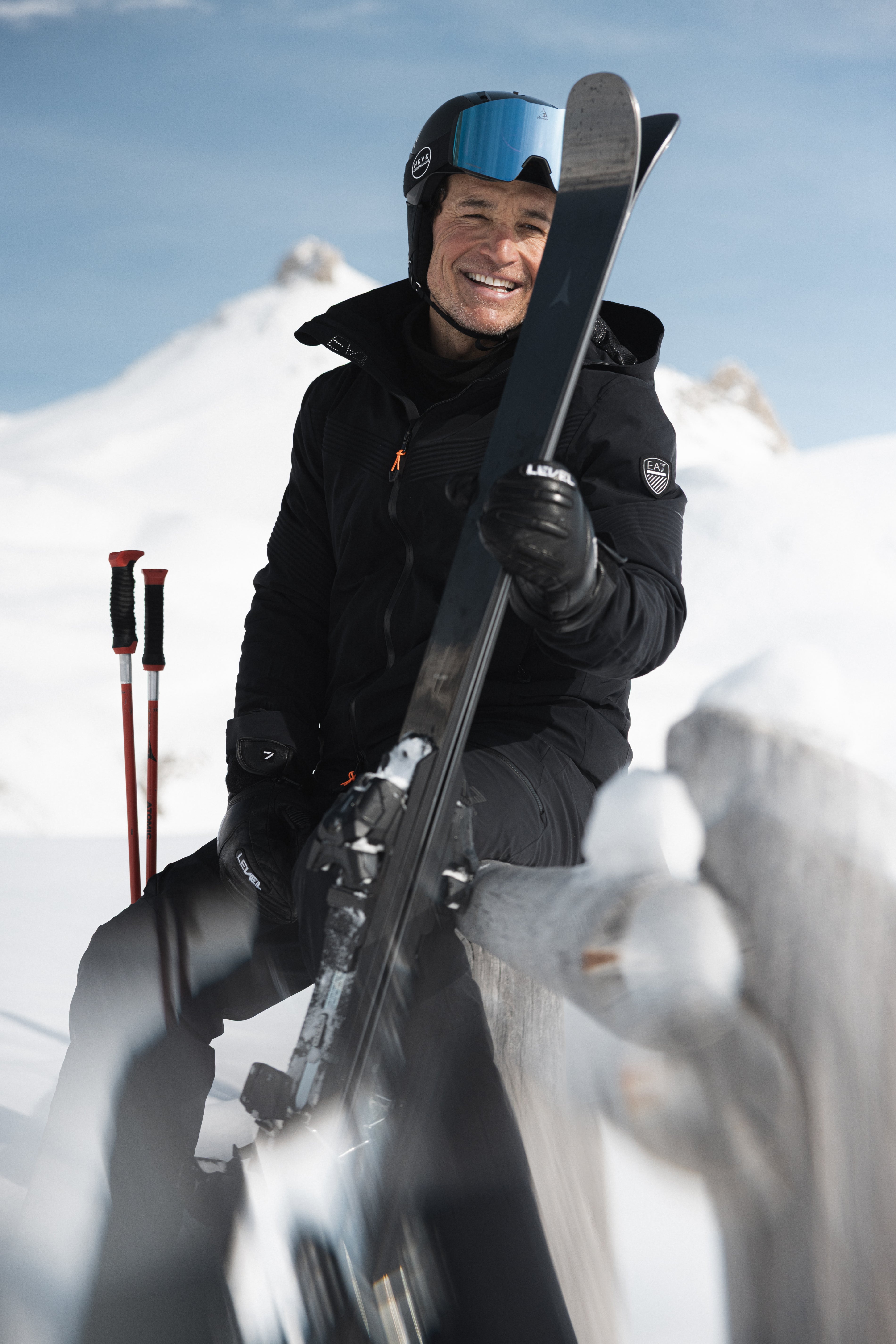 A smiley Giorgio Rocca in a black ski jacket and helmet with blue-tinted goggles sits in the snow holding a pair of skis, with ski poles beside them against a snowy mountain background.