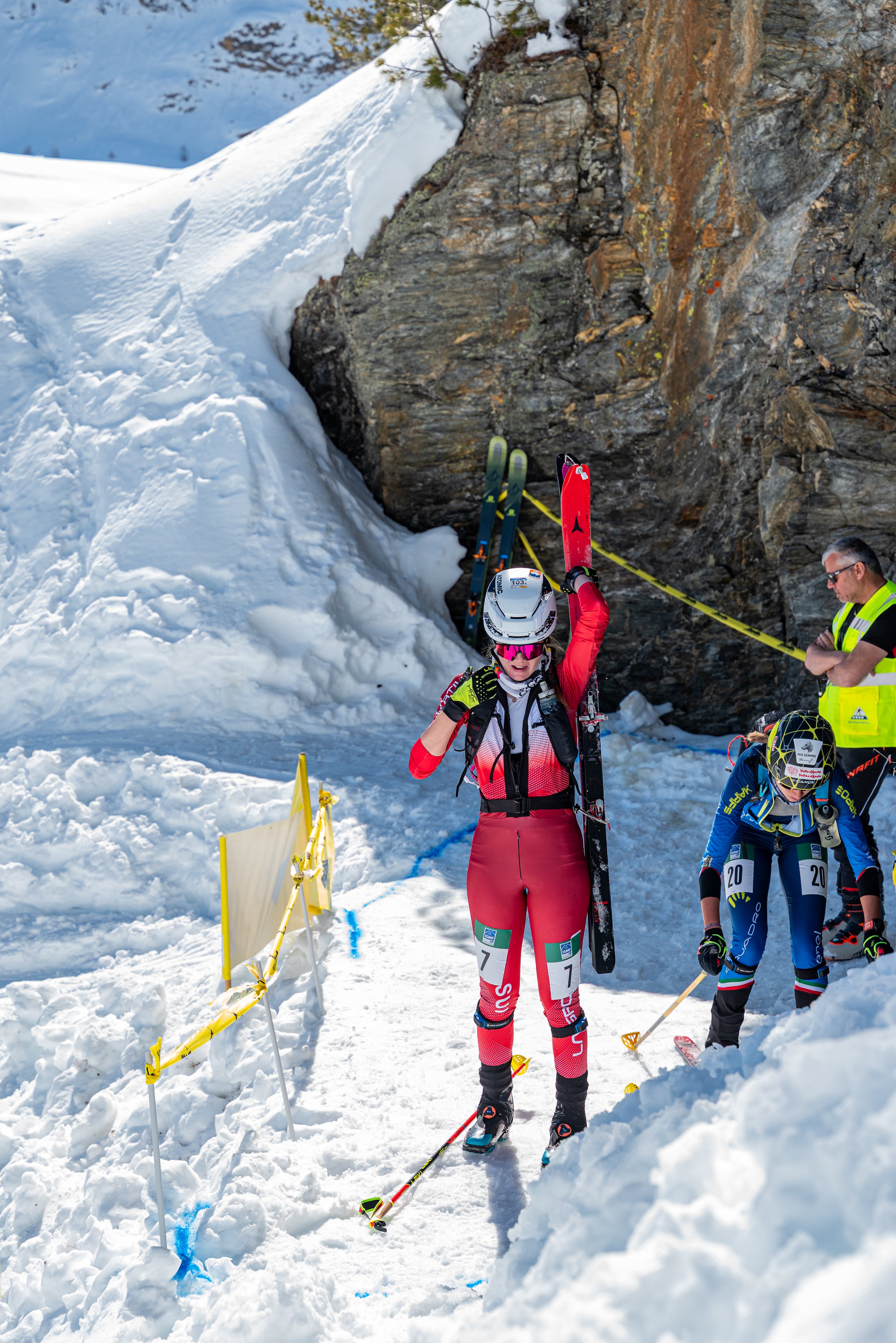 Alessandra Schmidt  in a red and black ski suit carries skis on their back while standing in deep snow near a rocky cliffside. Another skier in a blue suit and a person in a high-visibility vest are visible nearby in a snowy outdoor mountain setting.