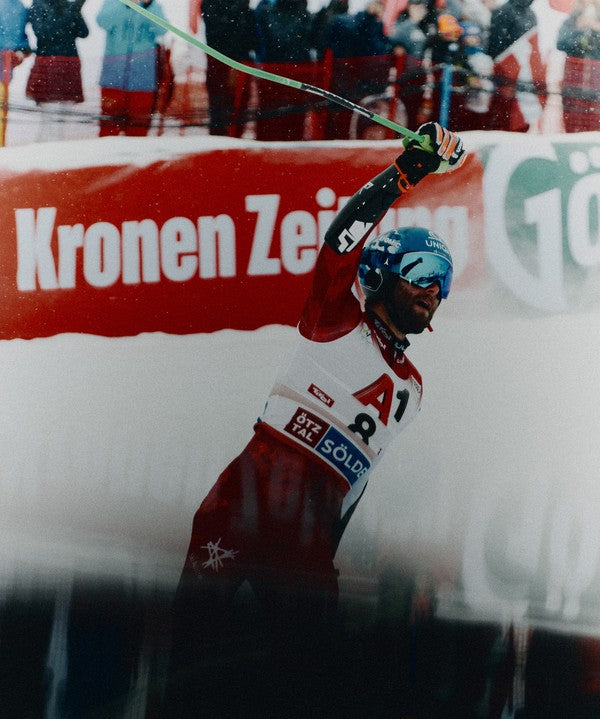 Marco Schwarz in a red and white ski suit raises their ski pole in celebration, with a red and white race barrier in the background.