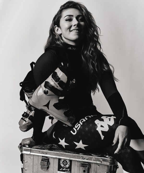 A black and white portrait of a Mikaela Shiffrin smiling in ski gear, 
 sitting on a metal case against a plain studio background.