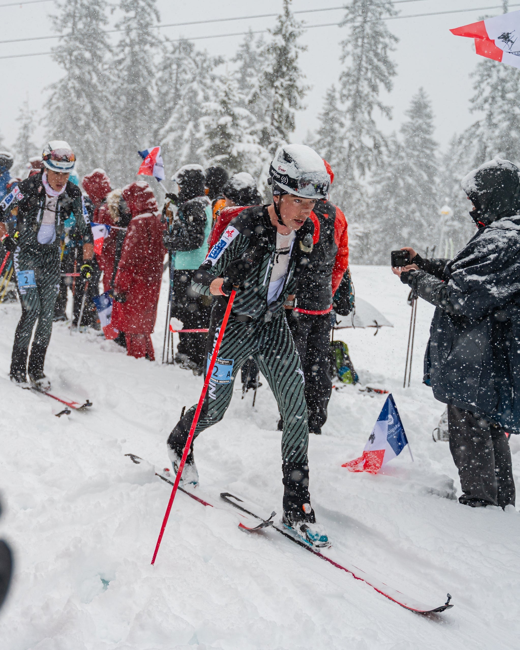 Julian Tritscher in a snowy outdoor winter scene, wearing striped ski pants and a white helmet, with other skiers and spectators around them in the snow.