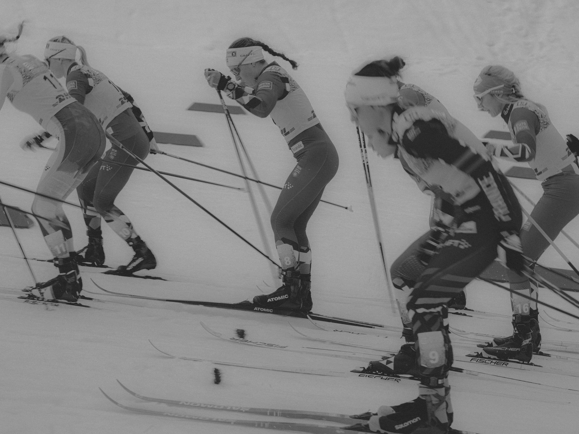 Black and white action photograph of multiple cross-country skier competitors racing side by side on a snowy track, captured with ski poles and skis visible.
