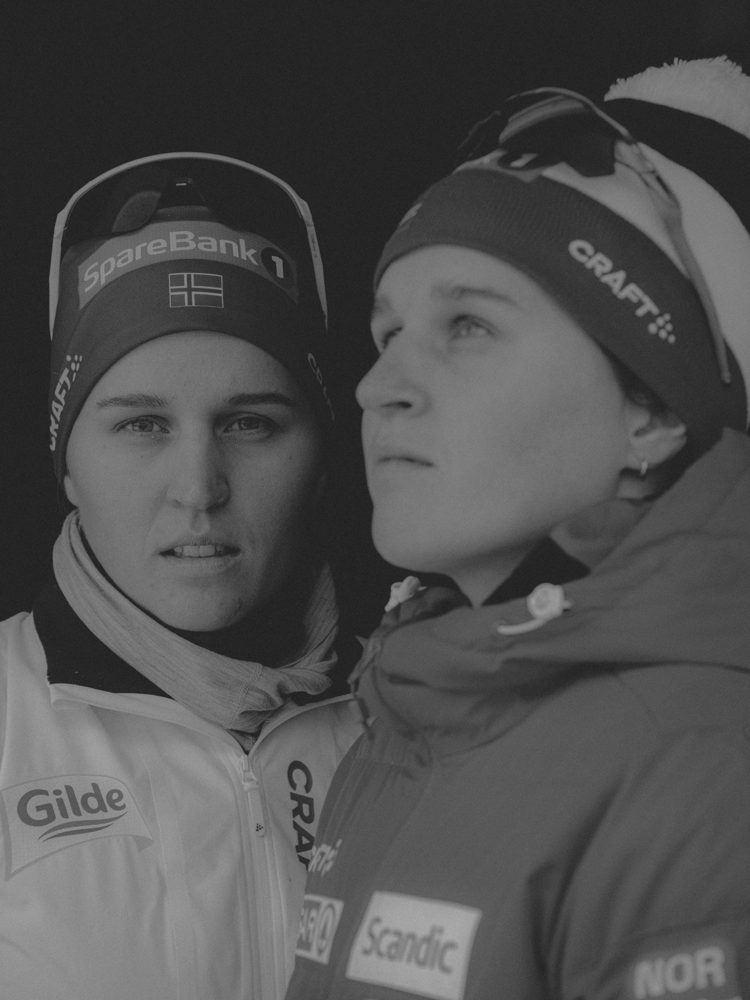 Black and white close-up of a mirrored Tiril Udnes Weng, wearing winter sports hats and jackets with sponsor logos.