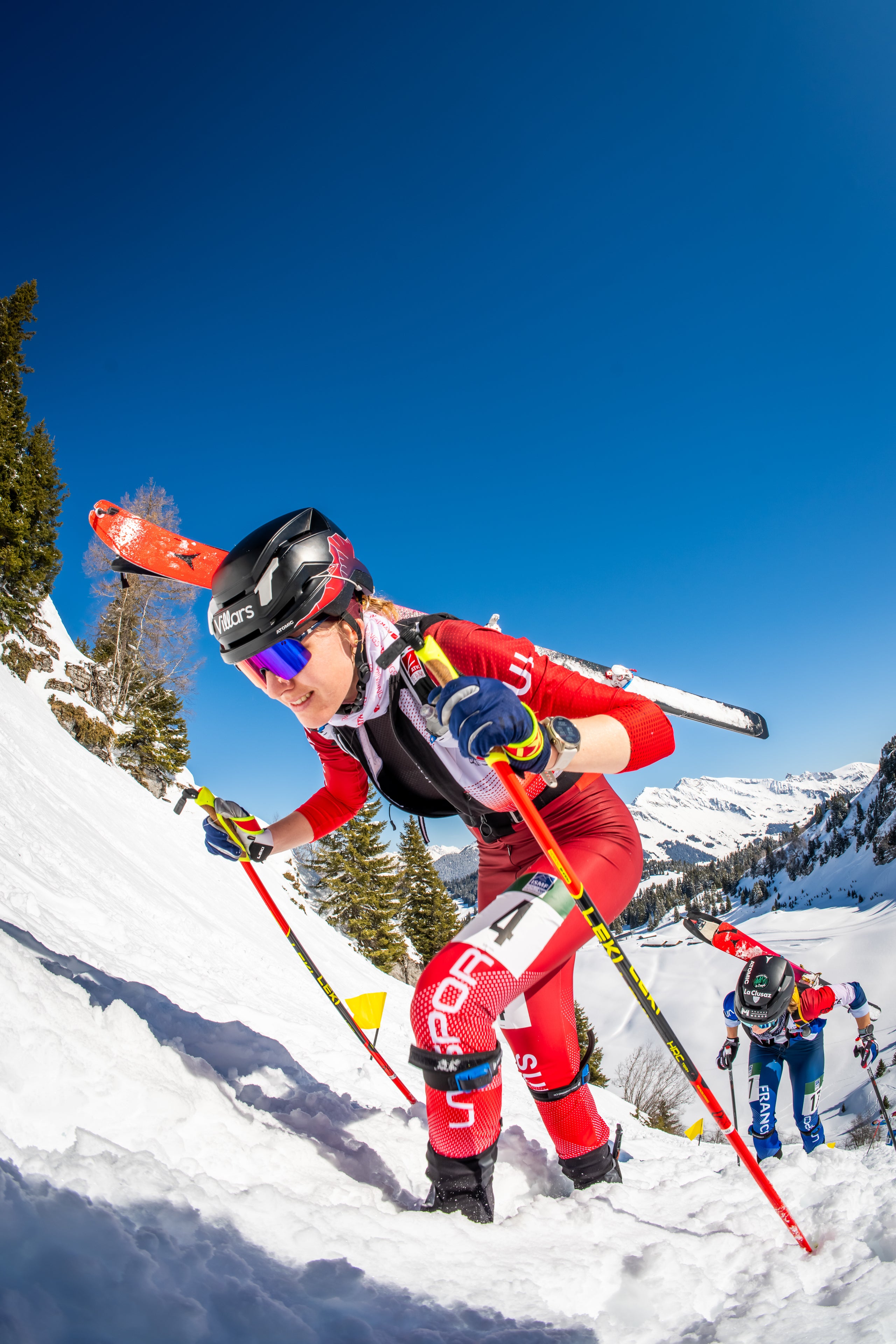Caroline Ulrich in a red and black ski suit, blue sunglasses, and a black helmet, leaning forward on ski poles while skiing down a snowy mountain slope under a clear blue sky.