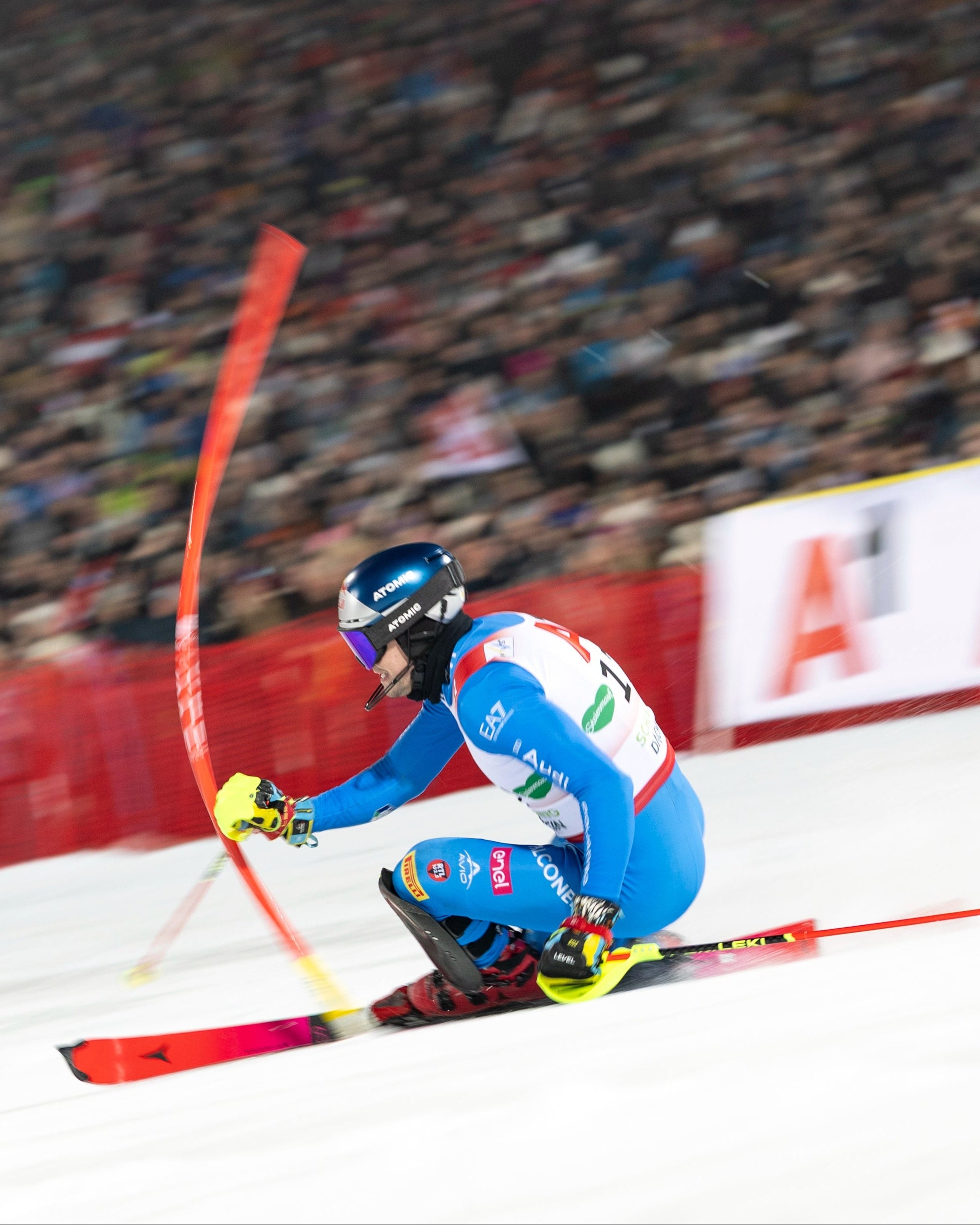 Alex Vinatzer in a blue and white racing suit skis through the snow during a race, holding red poles, with a blurred crowd in the background and a red safety barrier.