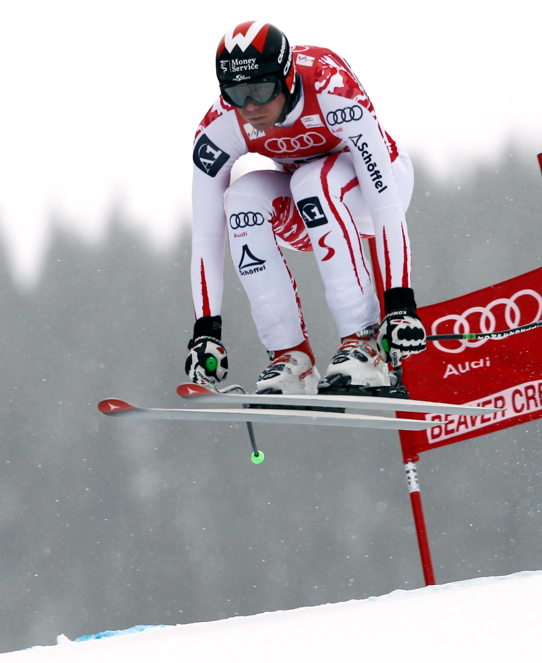 Michael Walchhofer in a red and white racing suit jumps over a red Beaver Creek slalom gate in the snow, with a blurred forest background.