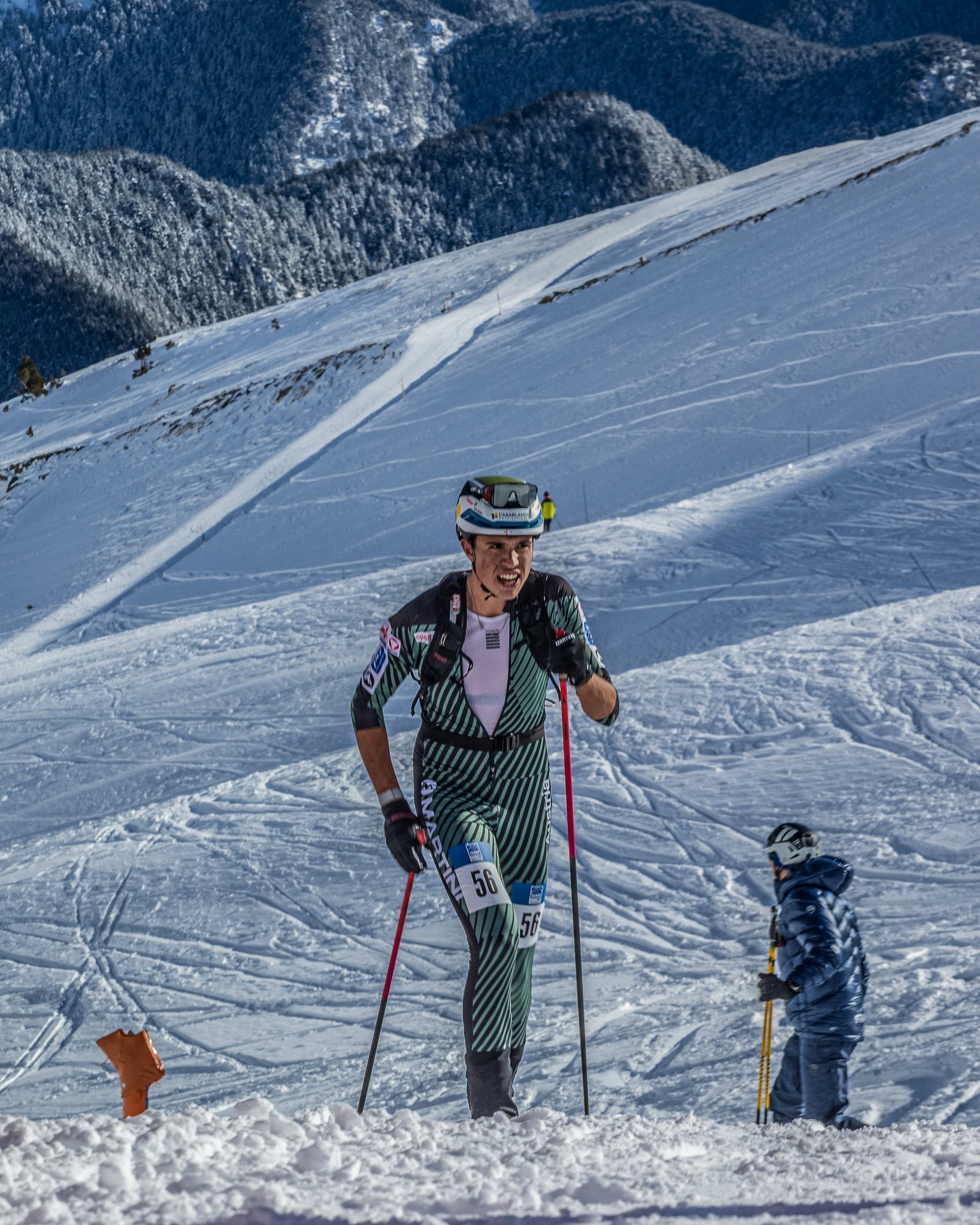 SIlvano Wolf in patterned ski pants and a helmet stands on a snowy mountain slope holding ski poles, and mountains in the background.