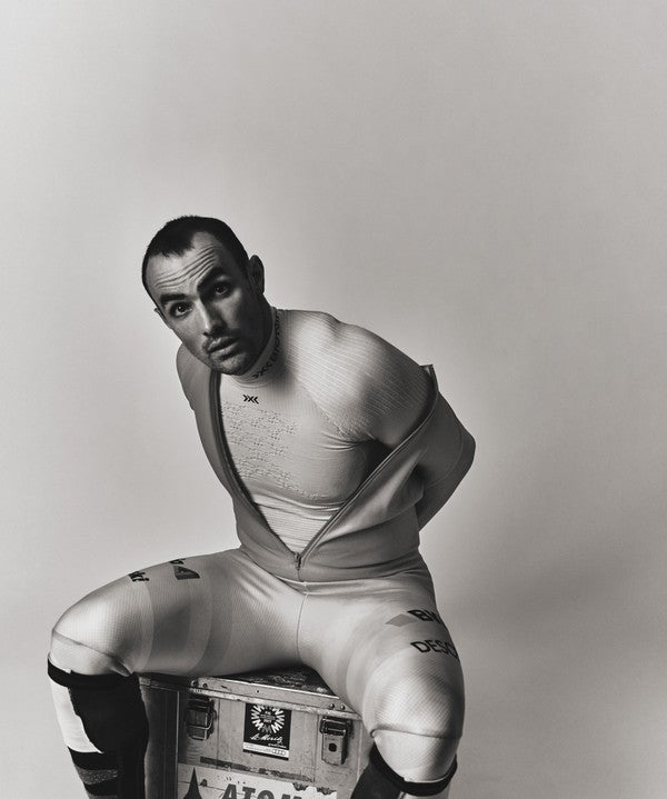 A black and white studio portrait of Daniel Yule wearing a tight, form-fitting athletic or costume suit, seated on a metal toolbox against a plain light background.