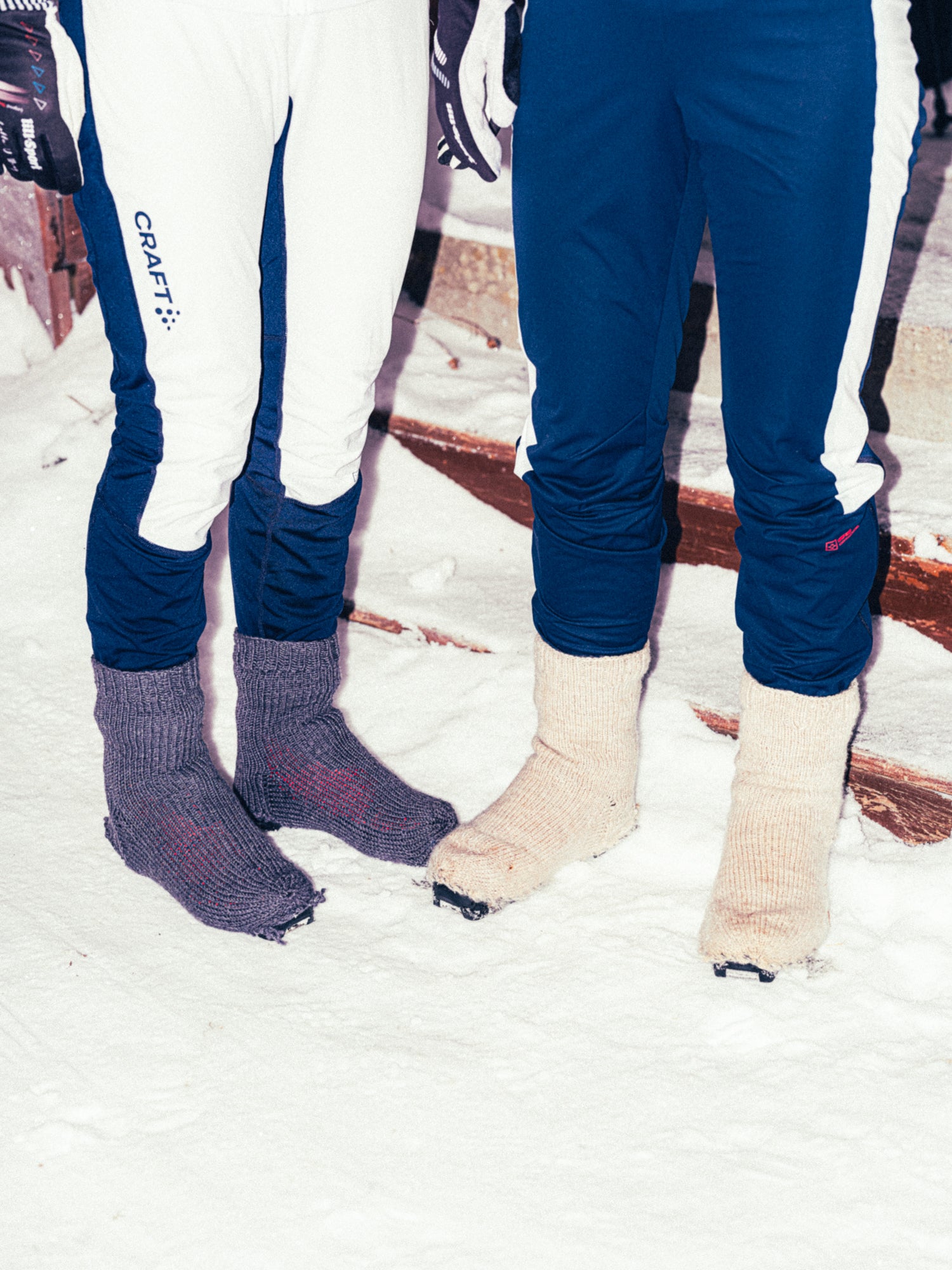 Two people standing in snow wearing ski pants with white and navy panels and knit boot covers, one in dark gray and one in cream.