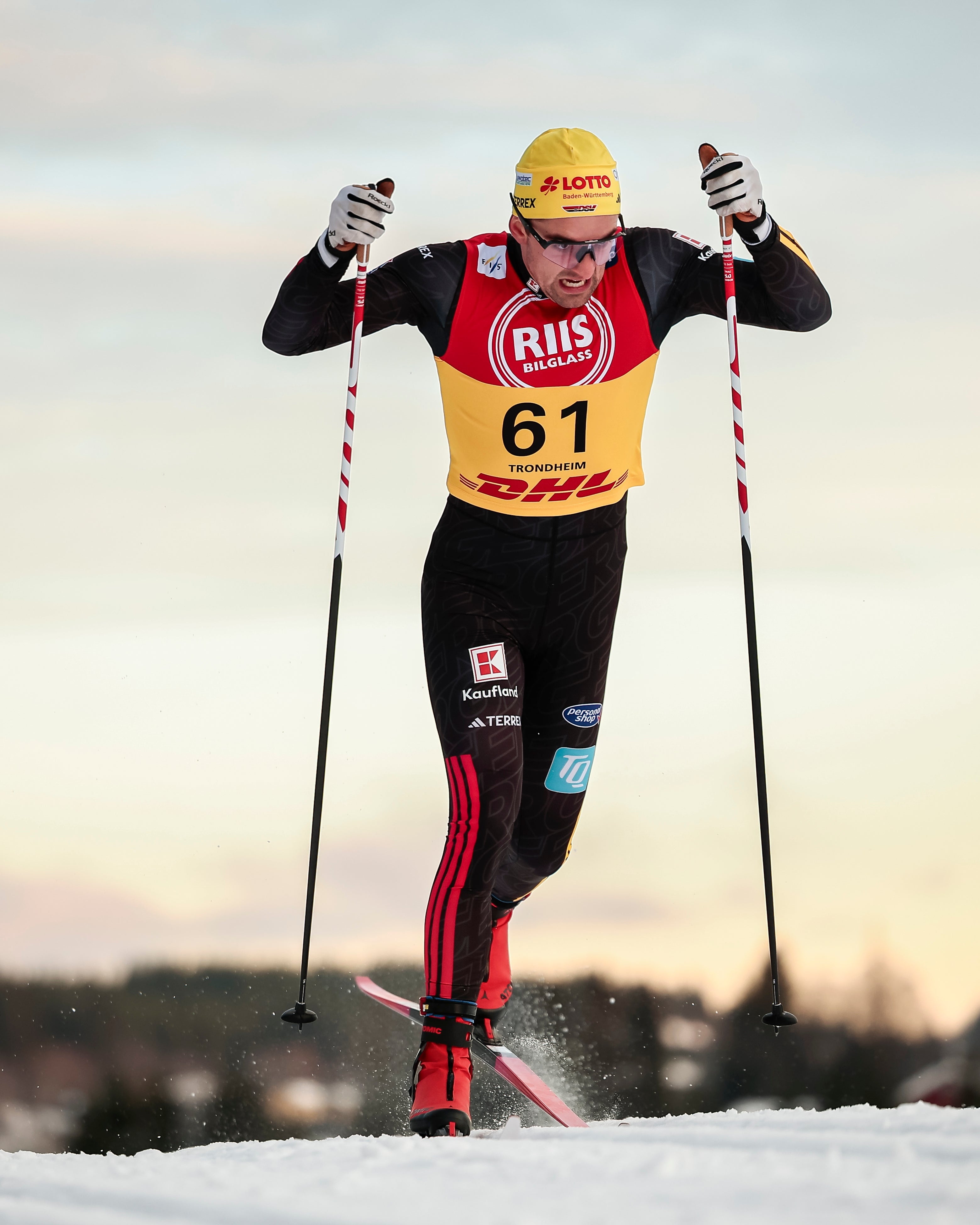 Janosch Brugger in a black, red, and yellow racing suit with DHL and RII logos, wearing a yellow helmet, skiing on a snowy track during a cloudy sunset.