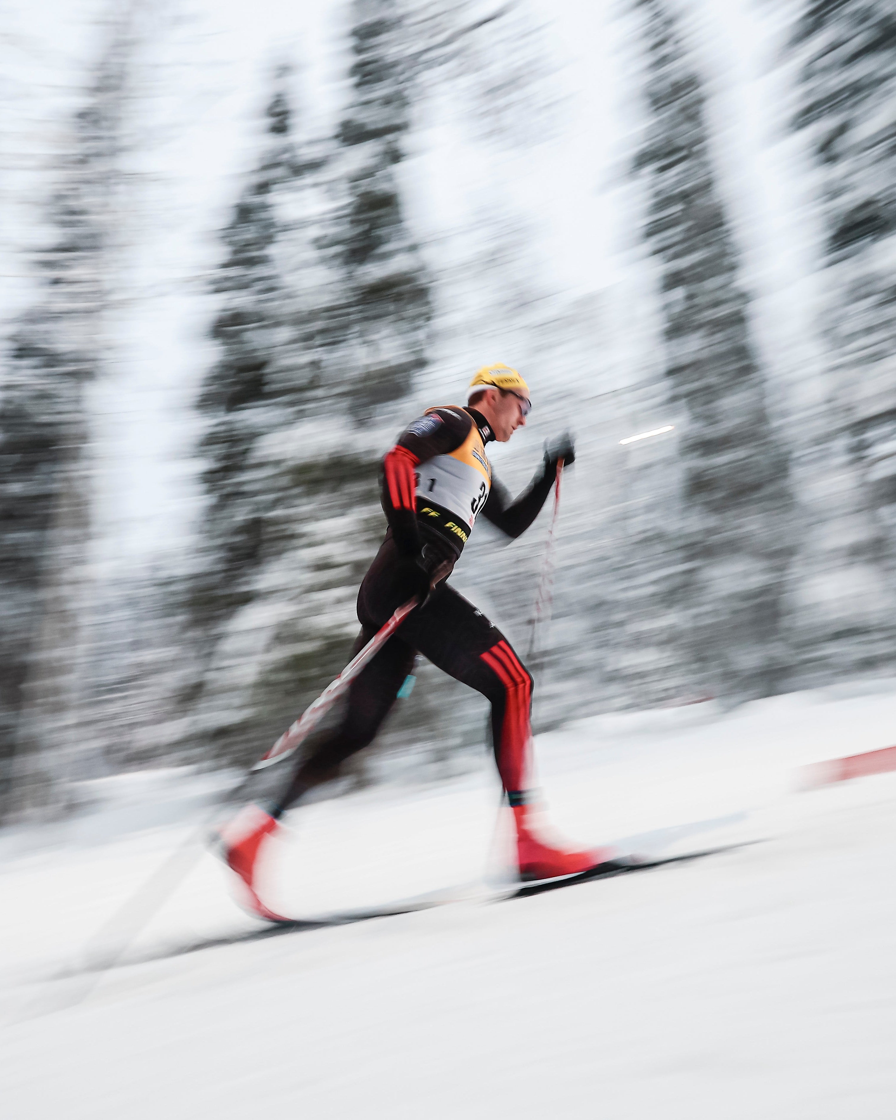 Janosch Brugger in a black and red racing suit with a yellow helmet gliding across a snowy forest track, captured with a motion-blur effect in the background.