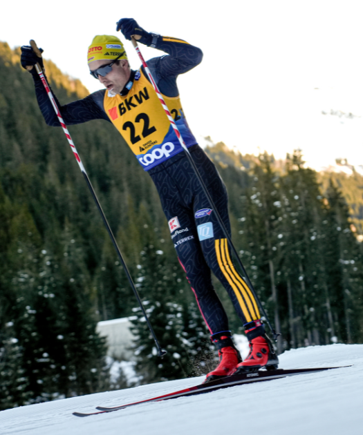 Janosch Brugger in a black, yellow, and blue racing suit with the number 22, wearing a yellow beanie and sunglasses, skiing on a snowy slope with a pine forest in the background.