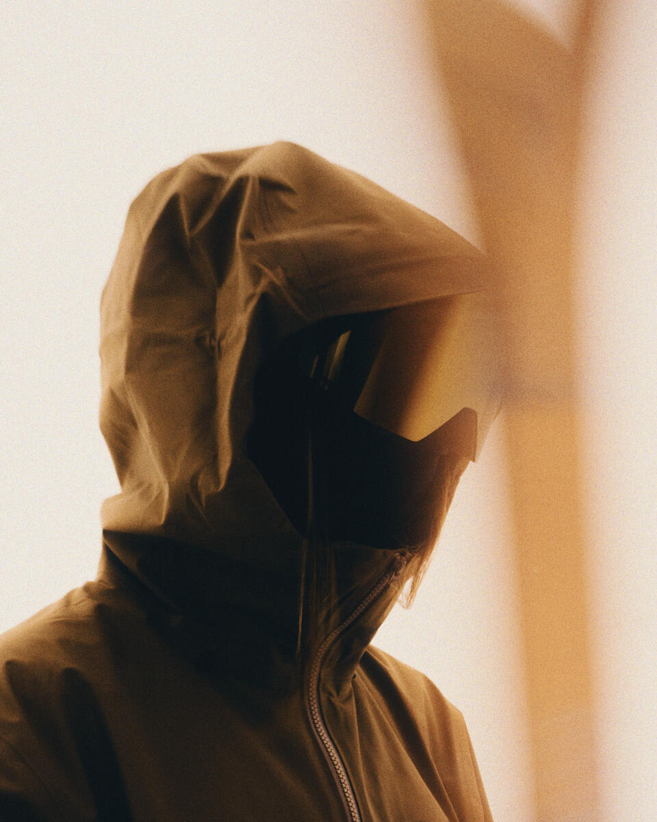 A person wearing a dark olive-brown hooded jacket with a reflective visor mask, shown in warm golden lighting against a soft beige background.