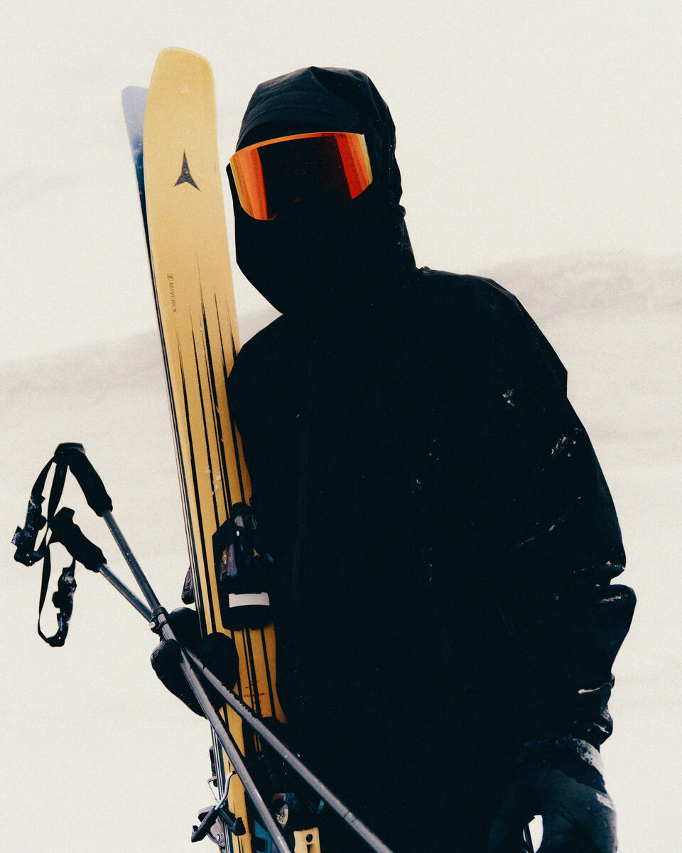 A person in a black ski jacket, black ski mask, and orange-tinted goggles stands in the snow holding a pair of yellow skis and ski poles.