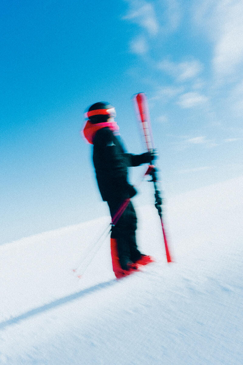 A skier in a black ski suit with bright red and orange accents stands on a snowy slope under a clear blue sky, holding red skis.