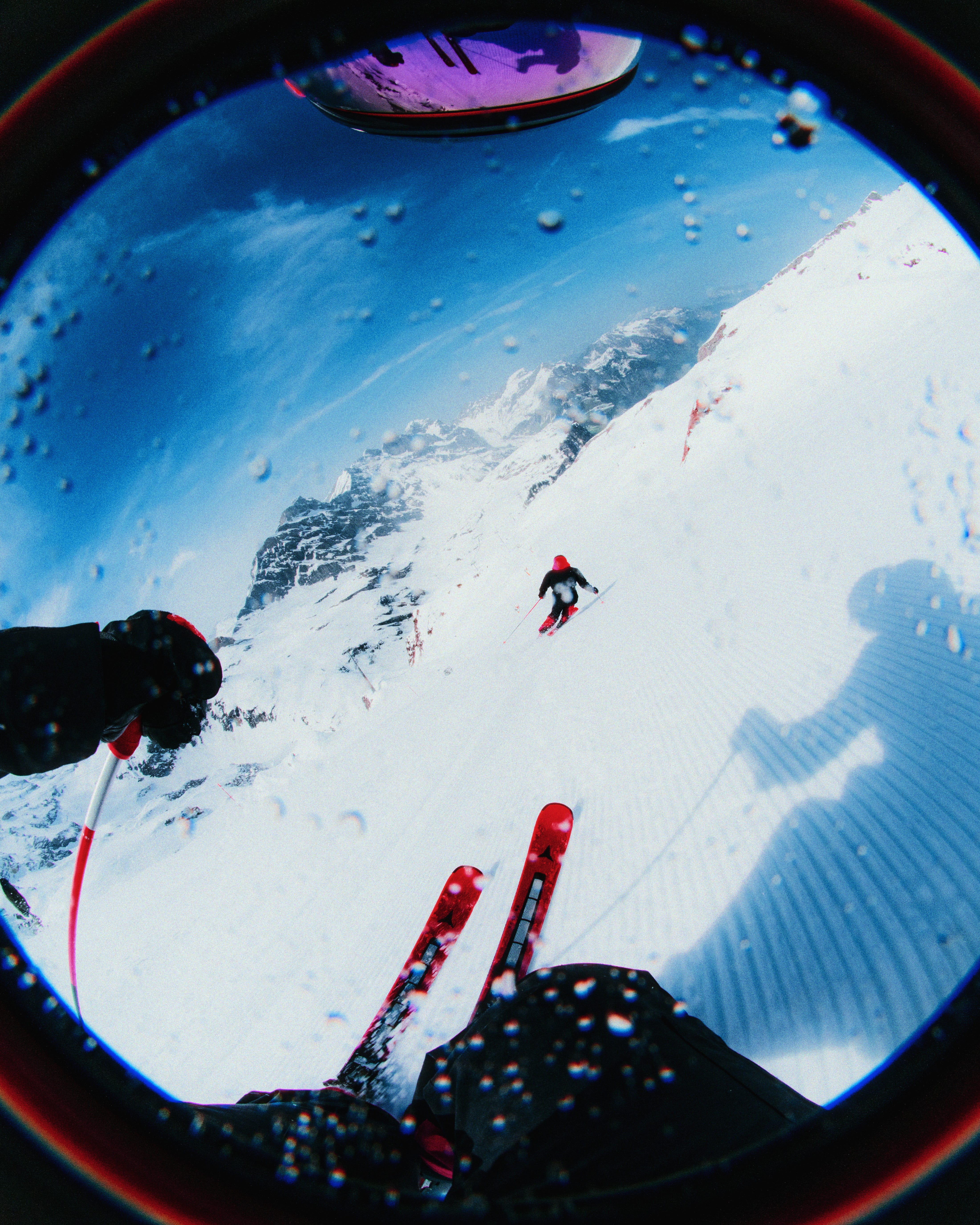 First-person view through a circular mirror or lens, showing a snowy mountain slope with a skier in the distance, blue sky, and the photographer's gloved hands and skis in the foreground.