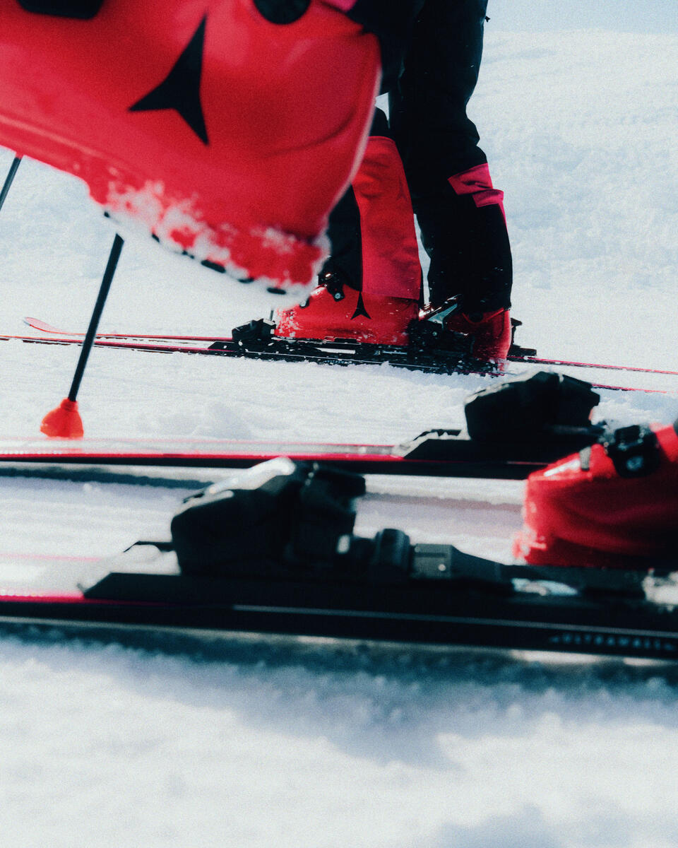 Close-up outdoor ski scene in snow with a person wearing black ski pants and bright red ski boots, with skis and poles visible.