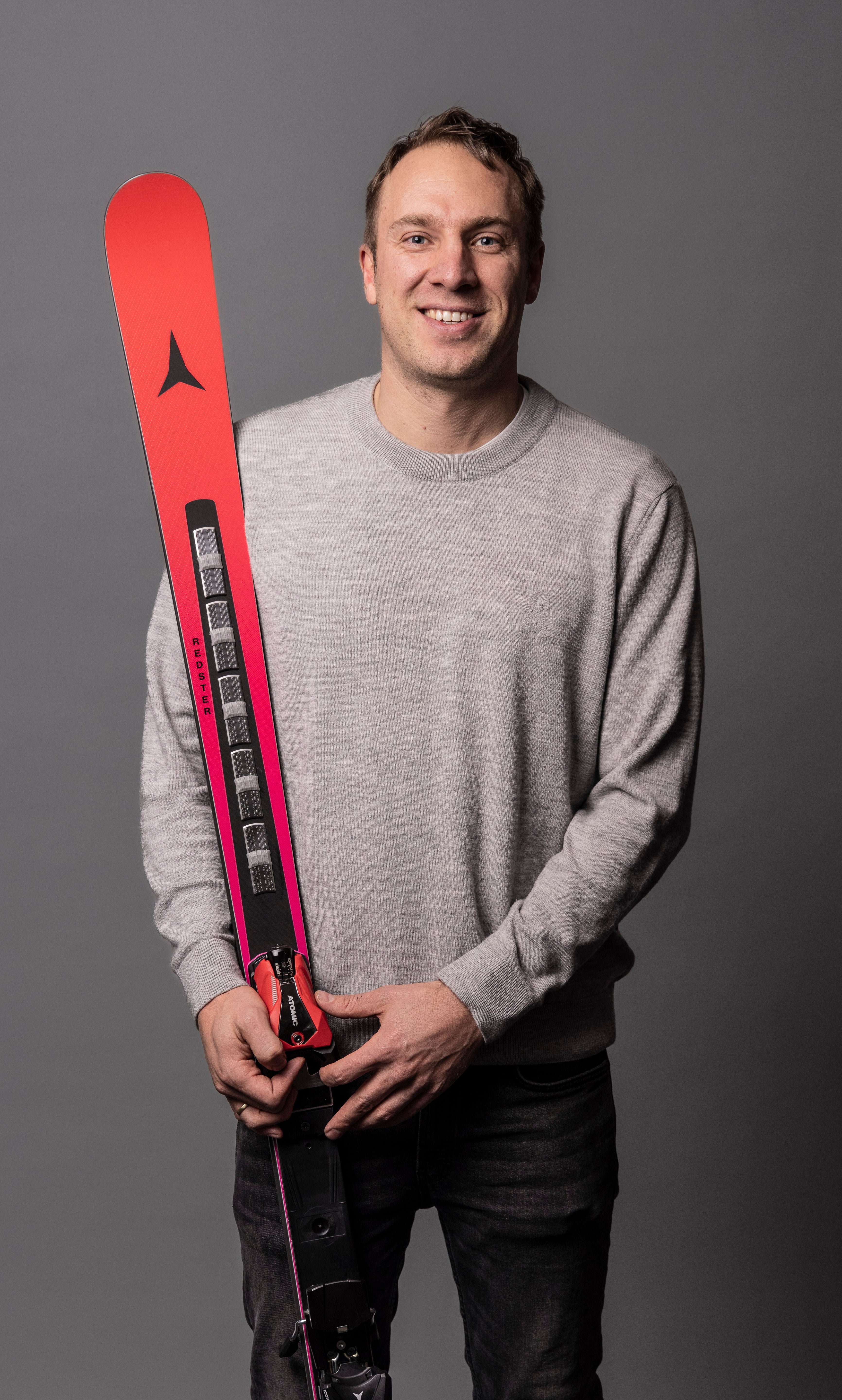 A man in a gray long-sleeve shirt and dark jeans holds a red and black ski with bindings attached against a solid dark gray studio background.