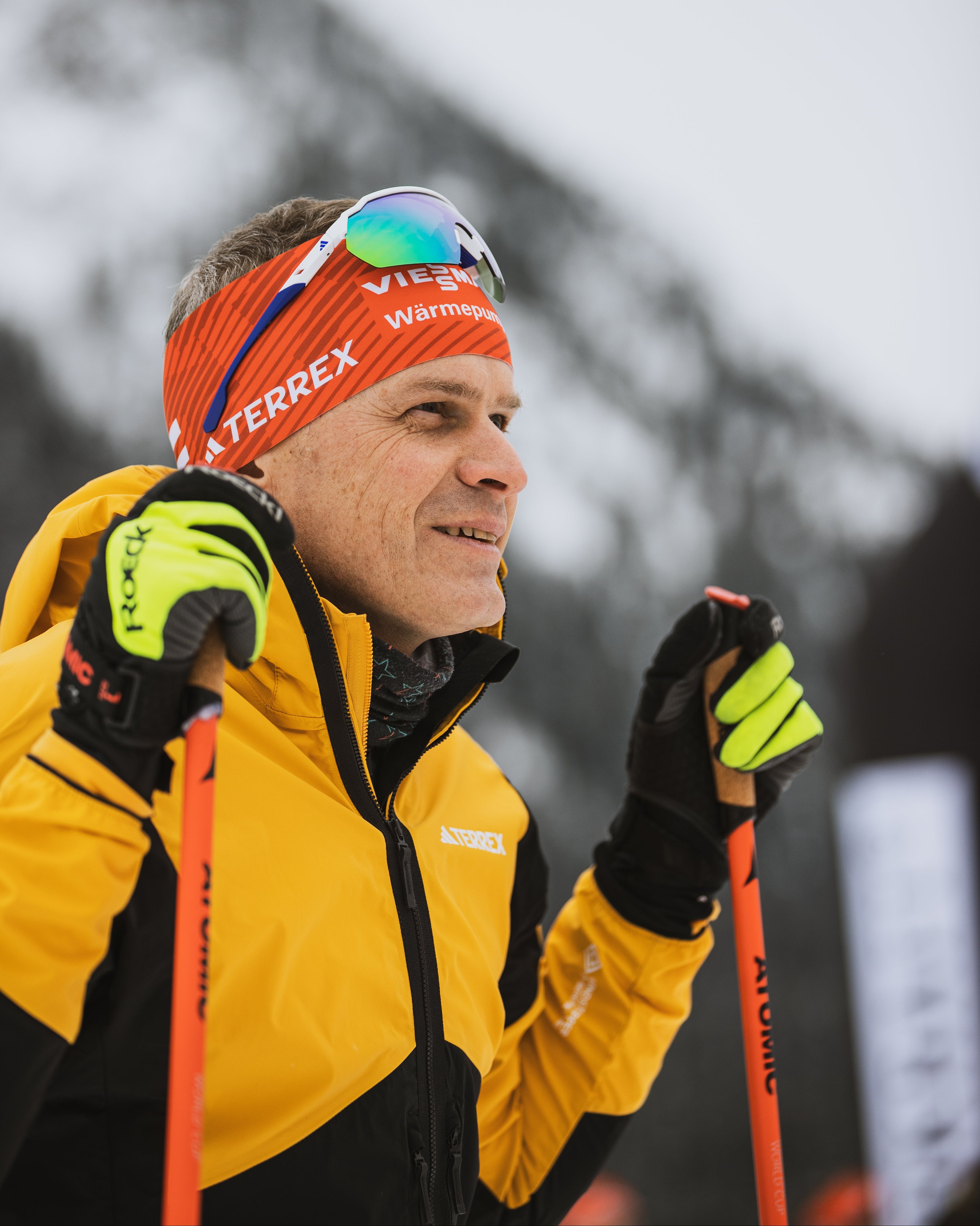 Tobias Angerer in winter sports gear, including a yellow and black jacket, an orange and black Terrex headband, gloves, and ski poles, captured outdoors against a snowy mountain background.