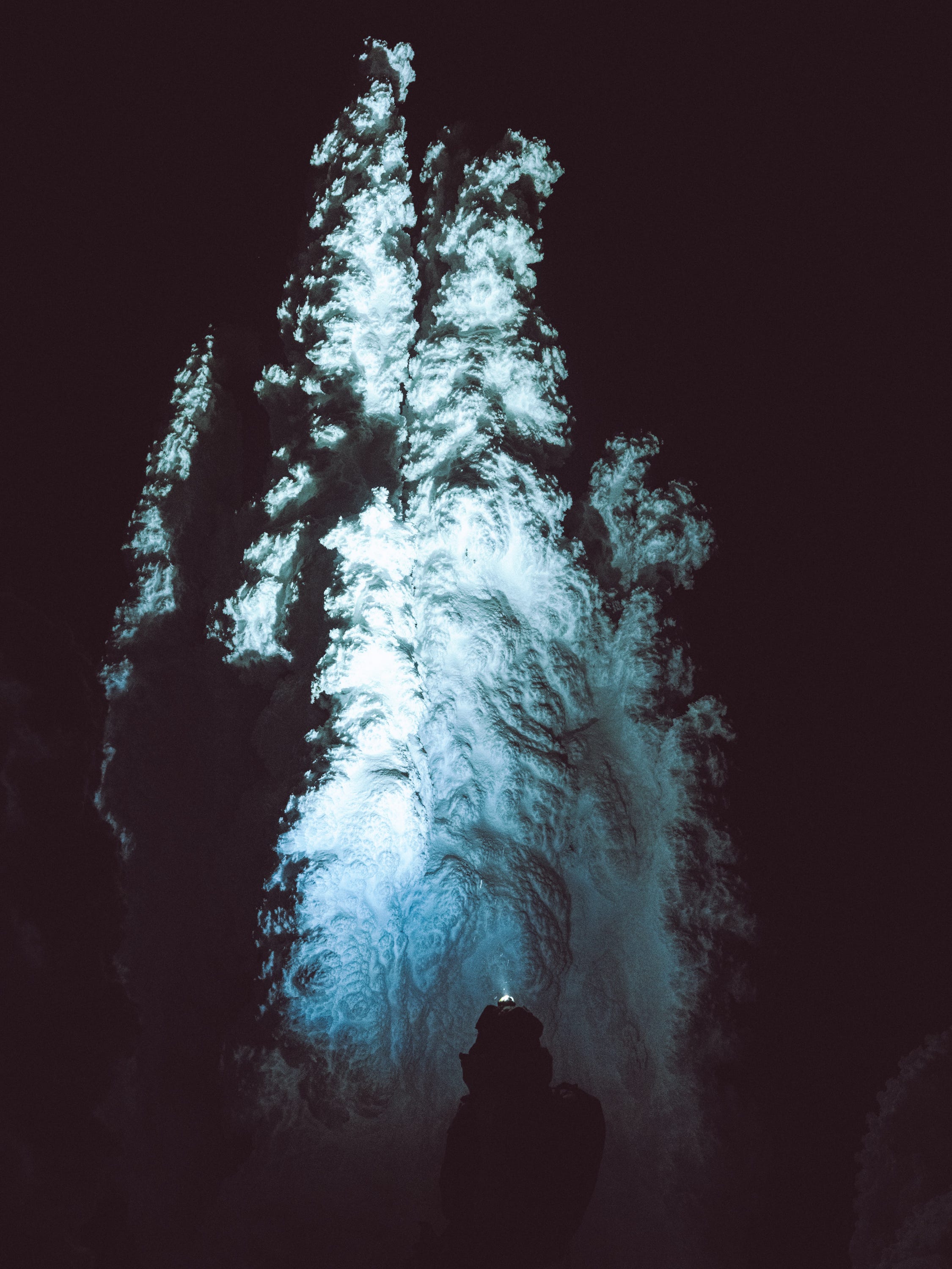 A dramatic night photograph of a glowing blue-white cave or waterfall opening, with a dark silhouette of a person standing underneath looking upward.