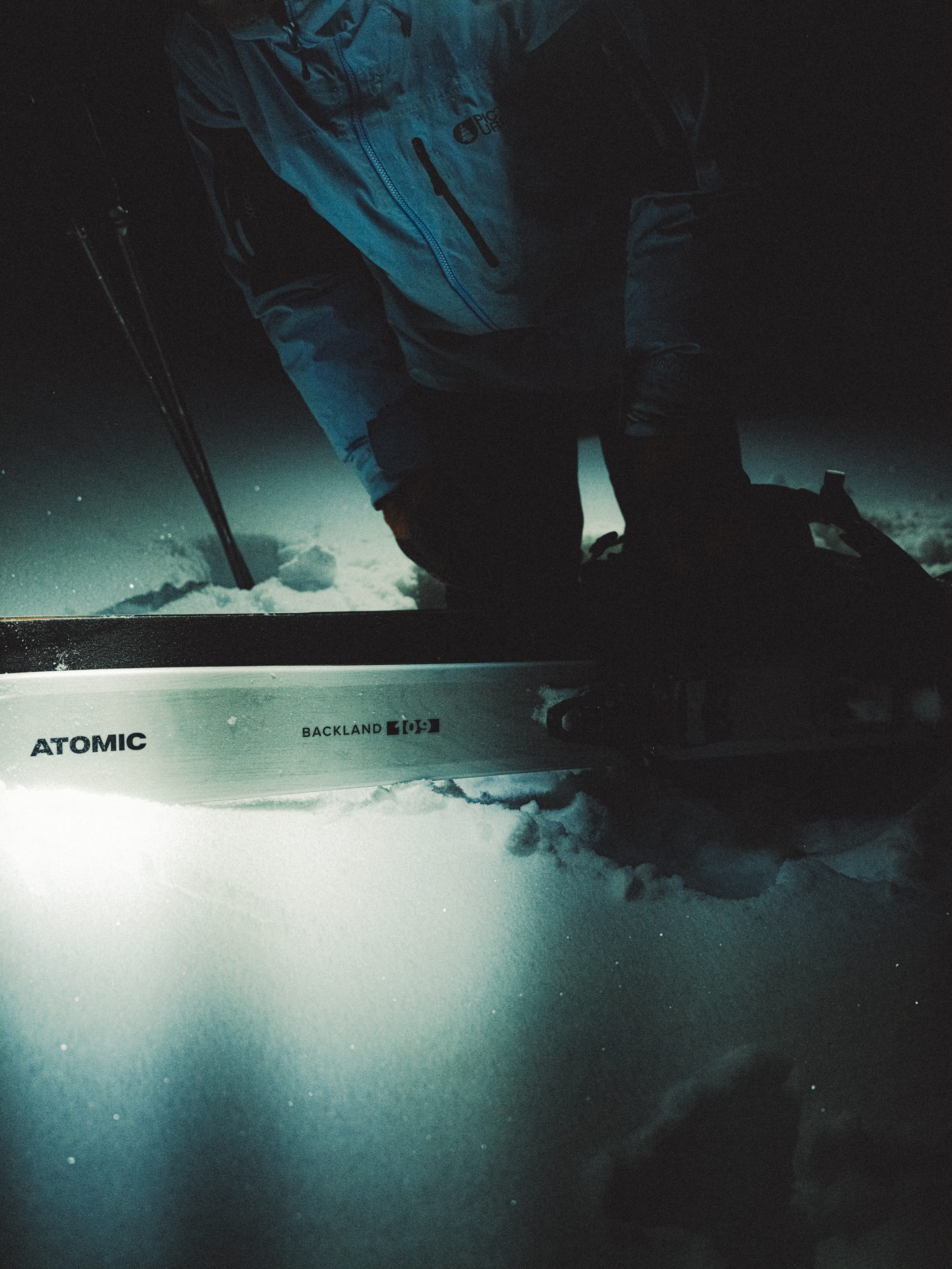 A person in dark clothing kneels in deep snow beside a ski, with the ski deck showing the brand name ATOMIC and model BACLAND 800-3.