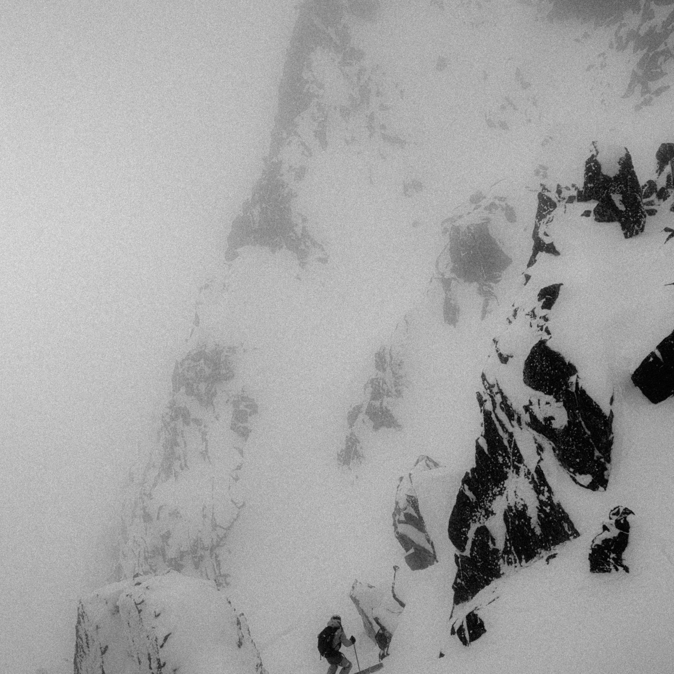 Black and white photograph of a snowy mountain slope with dark rocky outcrops and a small skier near the bottom center.
