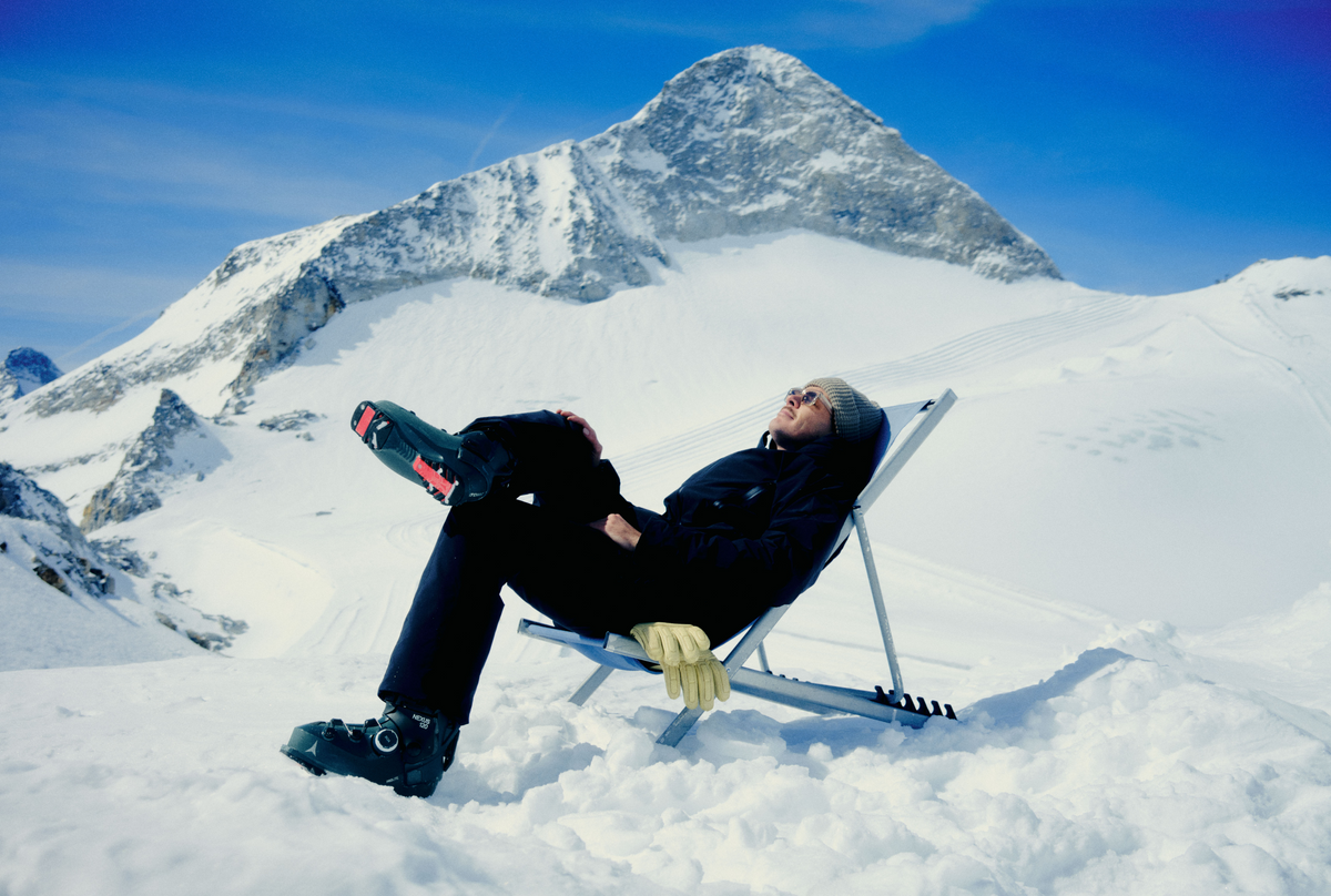 A person in black winter clothing reclines in a metal snow chair on a snowy mountain slope, with a large snow-covered peak and blue sky behind them.