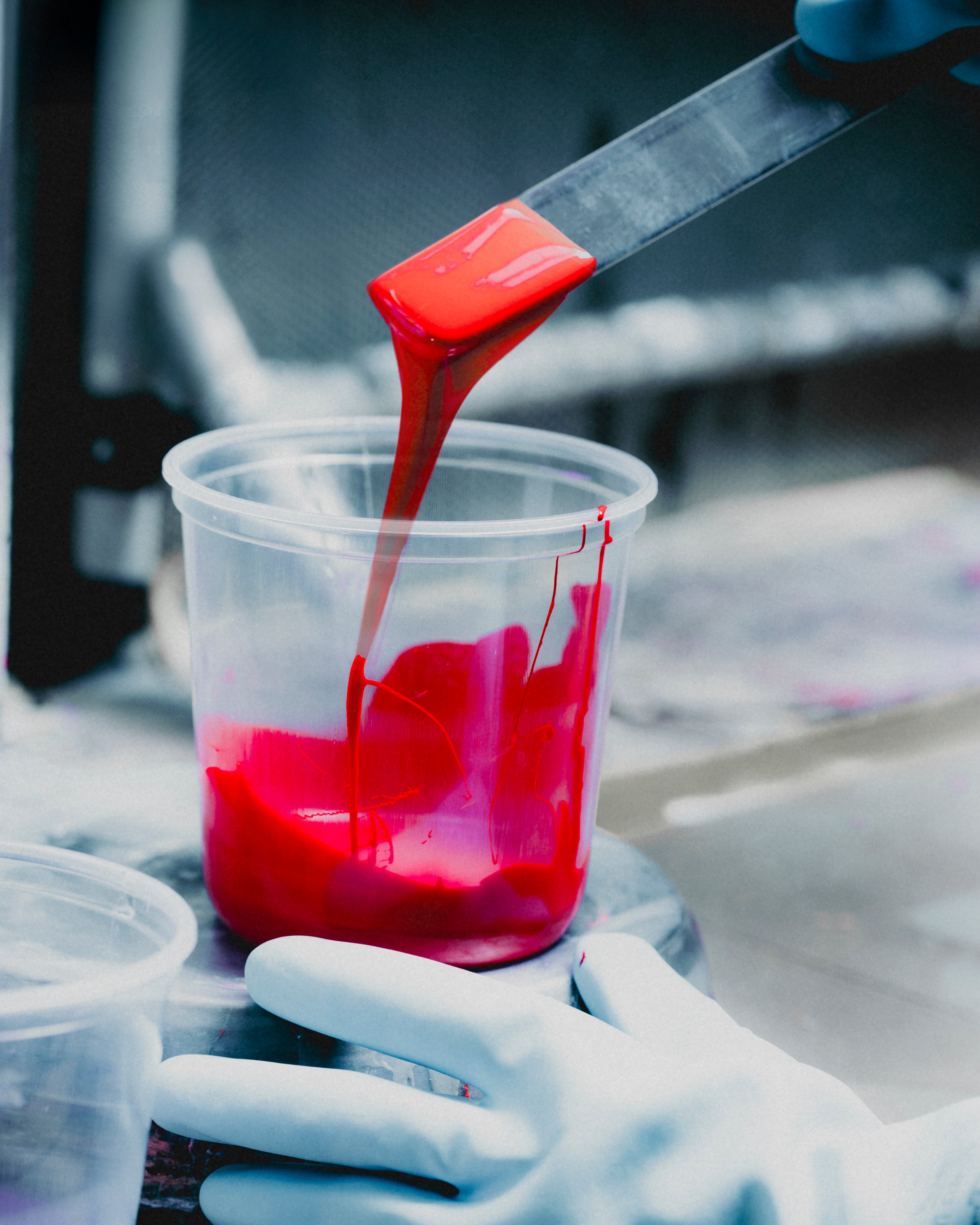 A gloved hand holds a clear plastic cup while a metal spatula pours bright red liquid into it. A second empty cup is partially visible in the foreground against a blurred gray background.