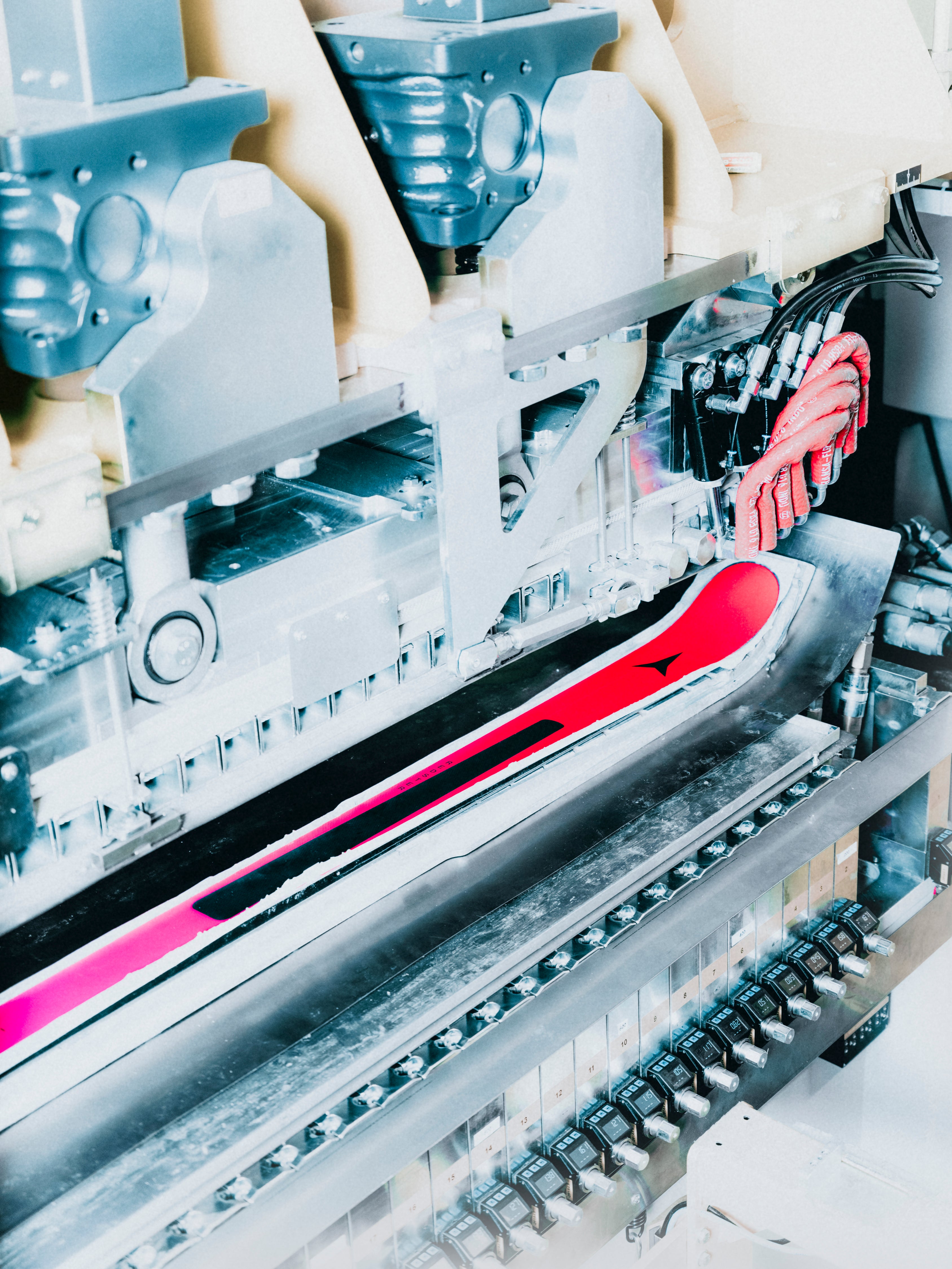 A close-up view of a ski being processed by an industrial machine, with the bright pink and black base of the ski clearly visible against metallic machinery.