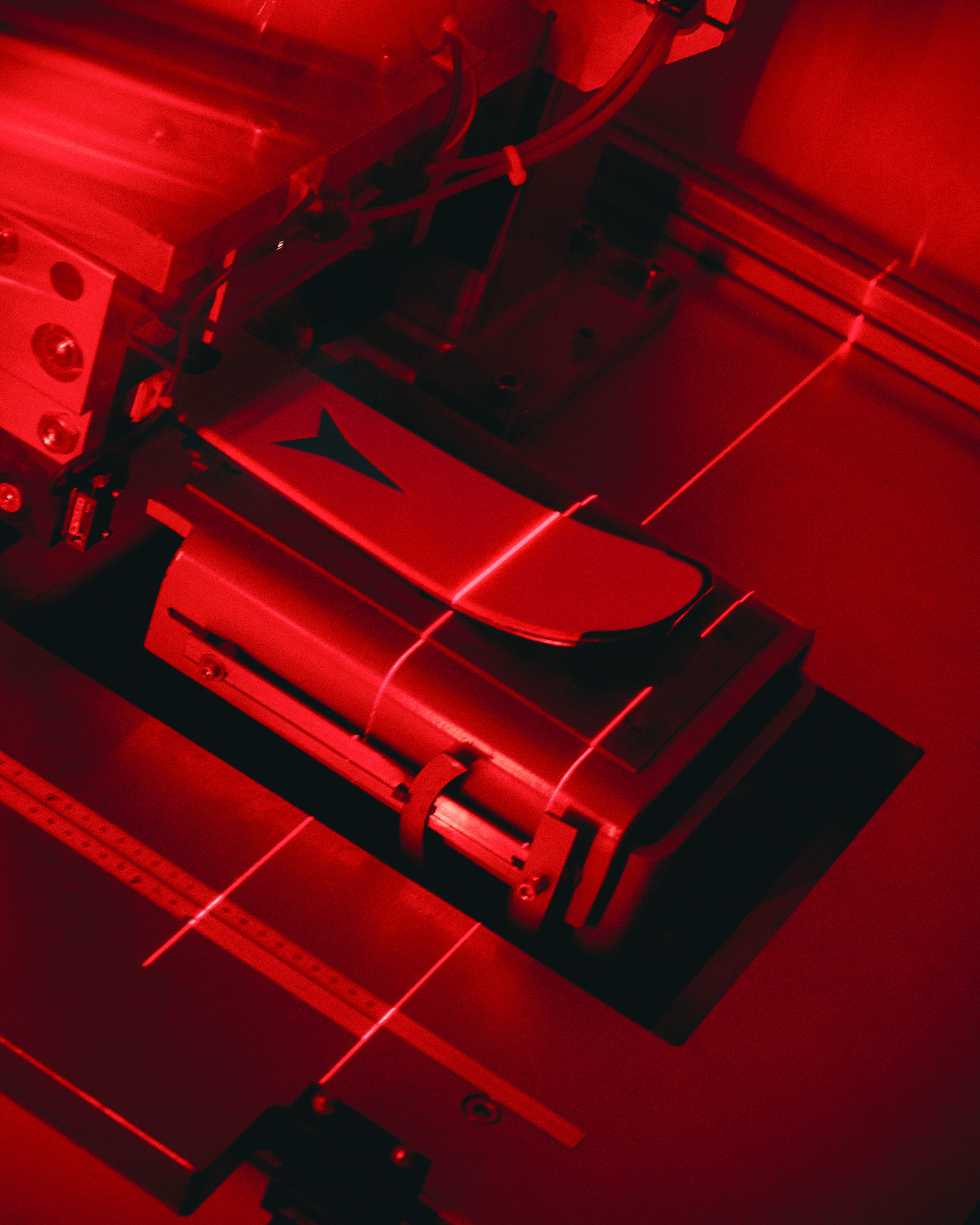 A close-up, dramatic shot of a metallic mechanical component or device under intense red lighting, with visible laser lines and a dark triangular logo on its surface.