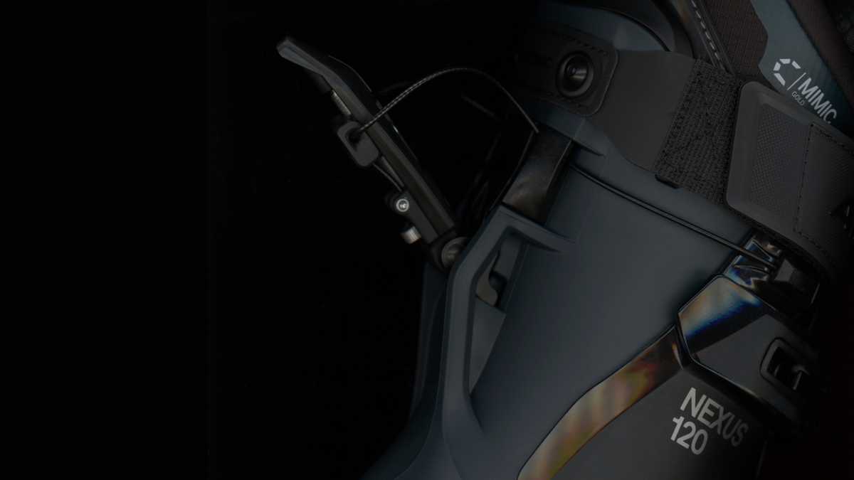 Close-up detail shot of a ski boot with visible buckles, straps, and branding. The boot is dark gray to black with metallic silver hardware and white printed text reading 'NEXUS 120' and 'IMMAC'.