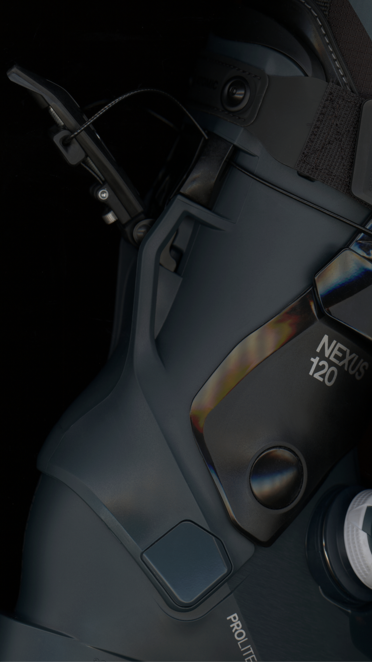 Close-up of a dark gray ski boot with visible branding including 'NEXUS 120' and 'PROLITE', showing the boot's textured shell, buckle area, and strap details against a dark background.