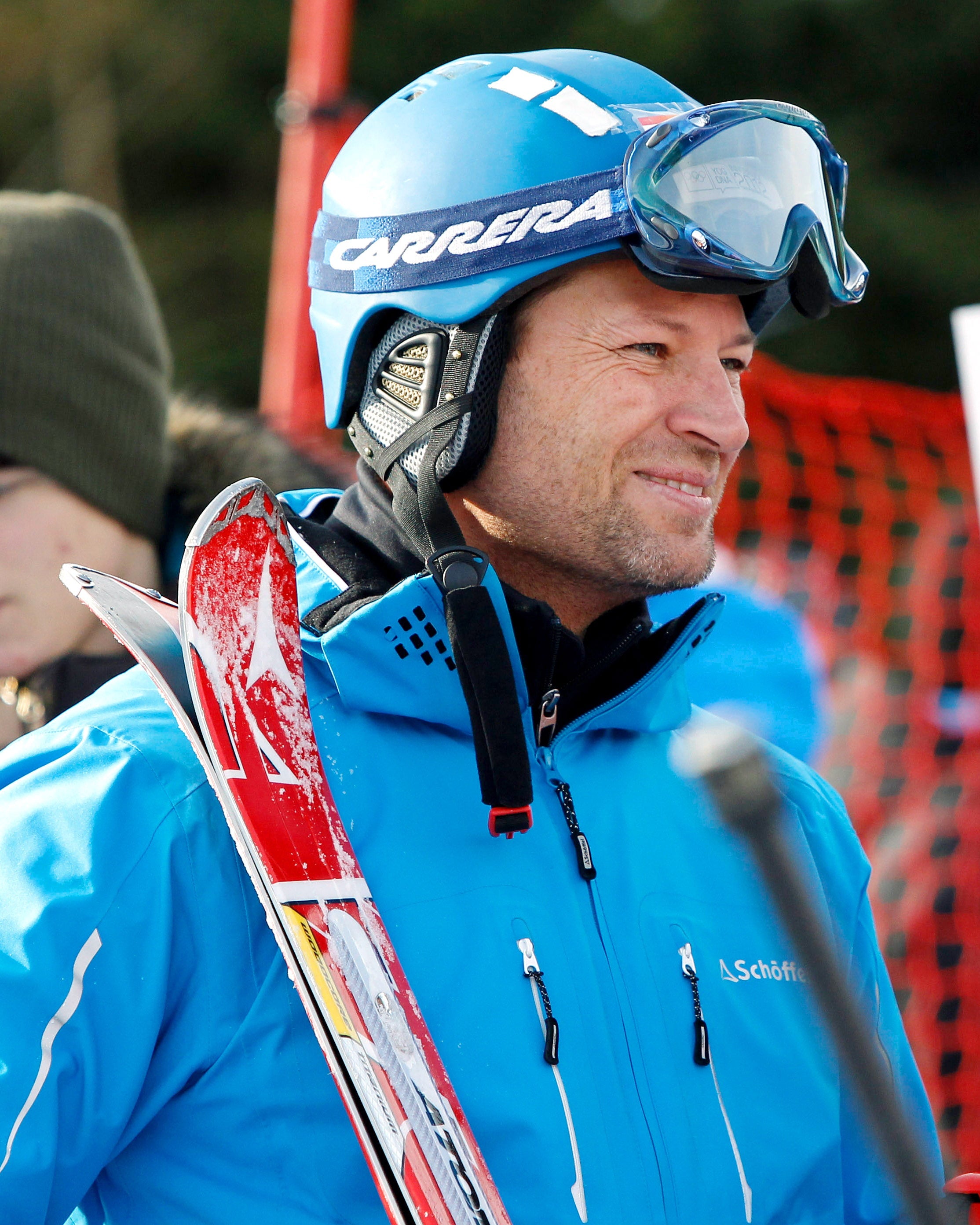 Stephan Eberharter in a blue Carrera ski helmet and bright blue ski jacket holding skis, smiling and talking with an interviewer holding an orange microphone at an outdoor ski event.