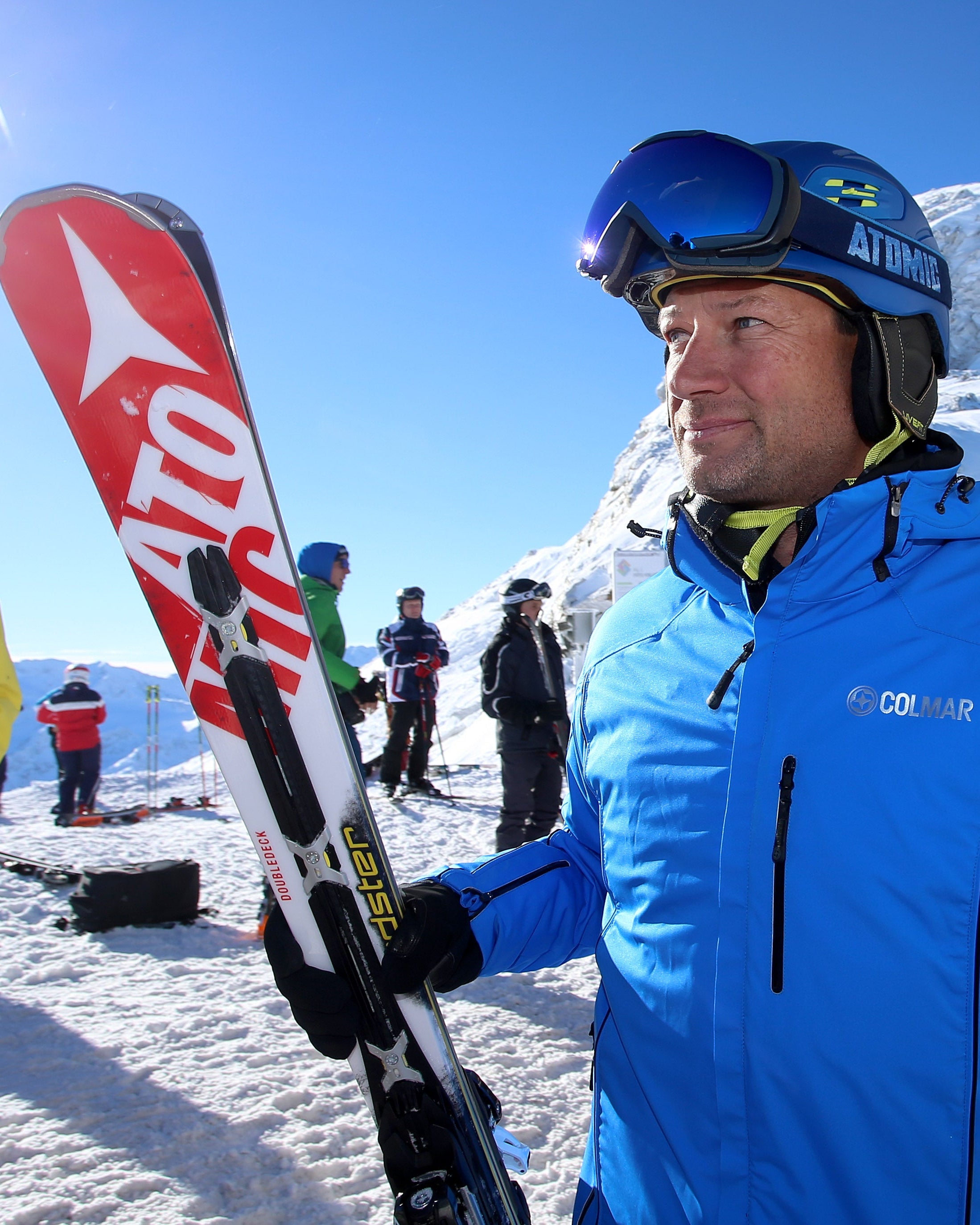 Stephan Eberharter in a blue COLUMBIA ski jacket and helmet with blue goggles holds red and white ski poles and red-and-white skis on a snowy mountain slope under bright sunlight.