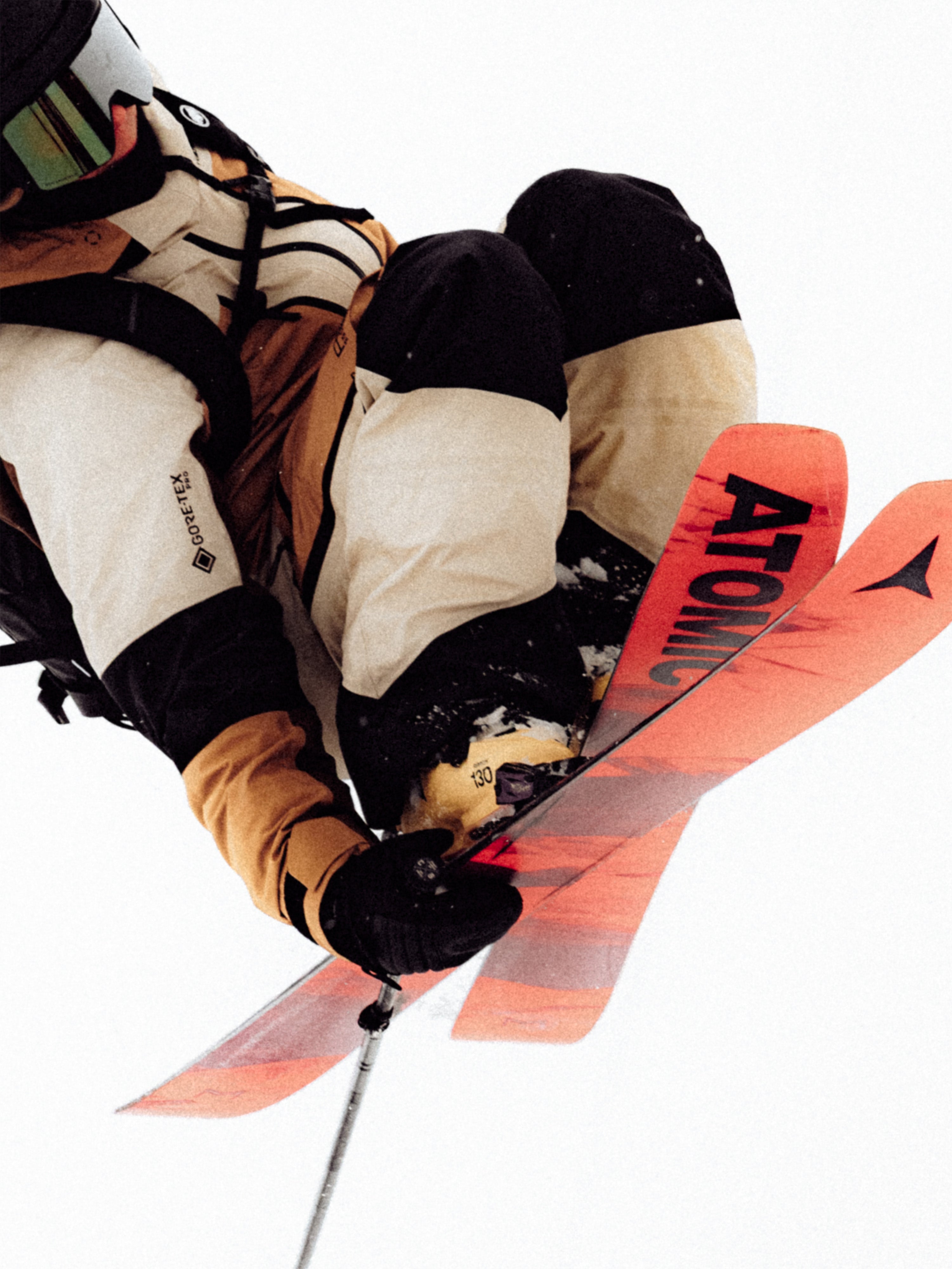 Justine Blanchard captured mid-air against a bright white background, wearing ski boots, gloves, and a ski suit. The skier is holding ski poles and has an Atomic ski visible under the boot, with the word 'ATOMIC' printed in black on a bright orange-red ski blade.
