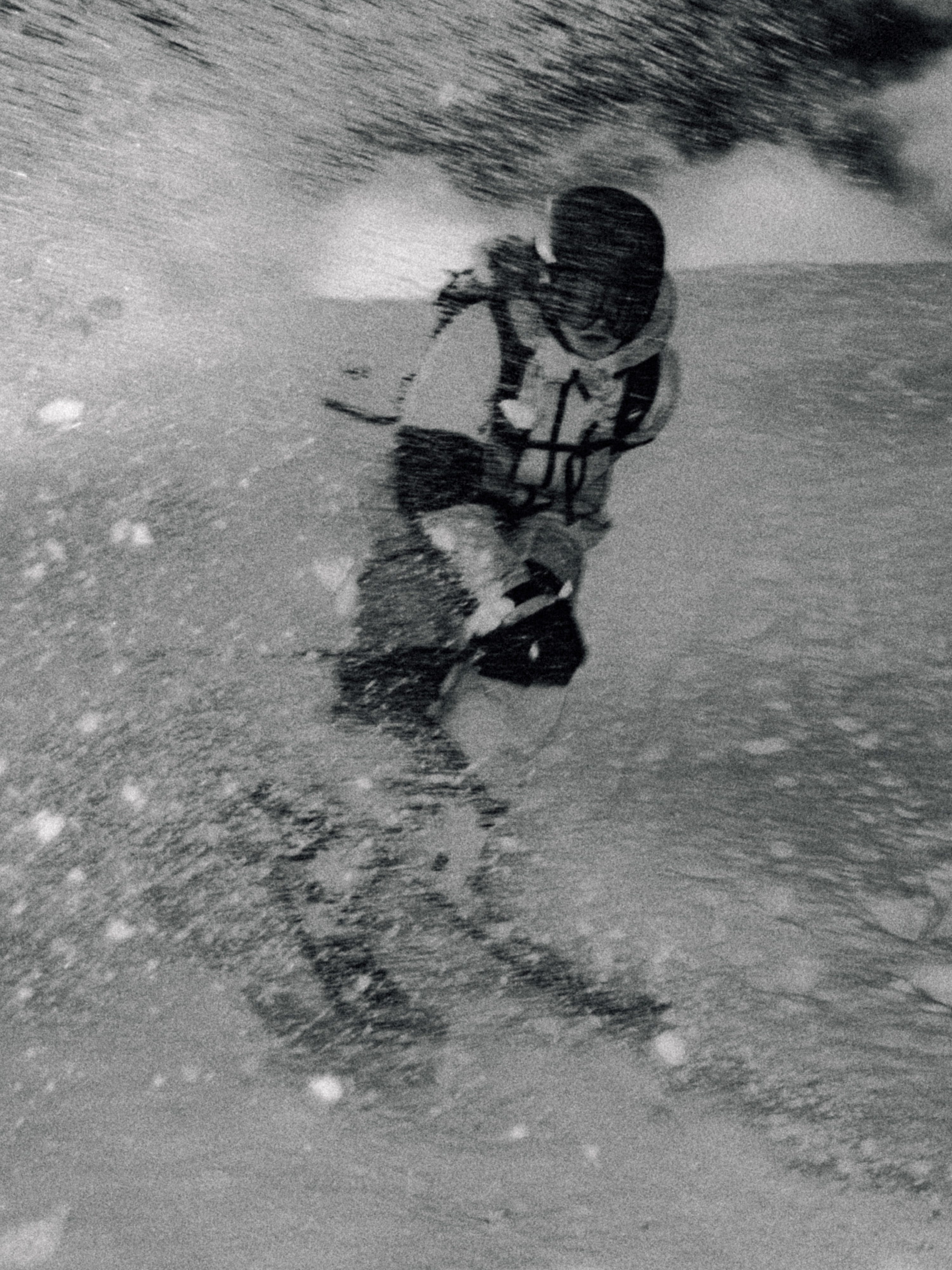 Black and white action photograph of a skier carving through heavy snow spray, wearing a helmet, goggles, and a ski jacket.